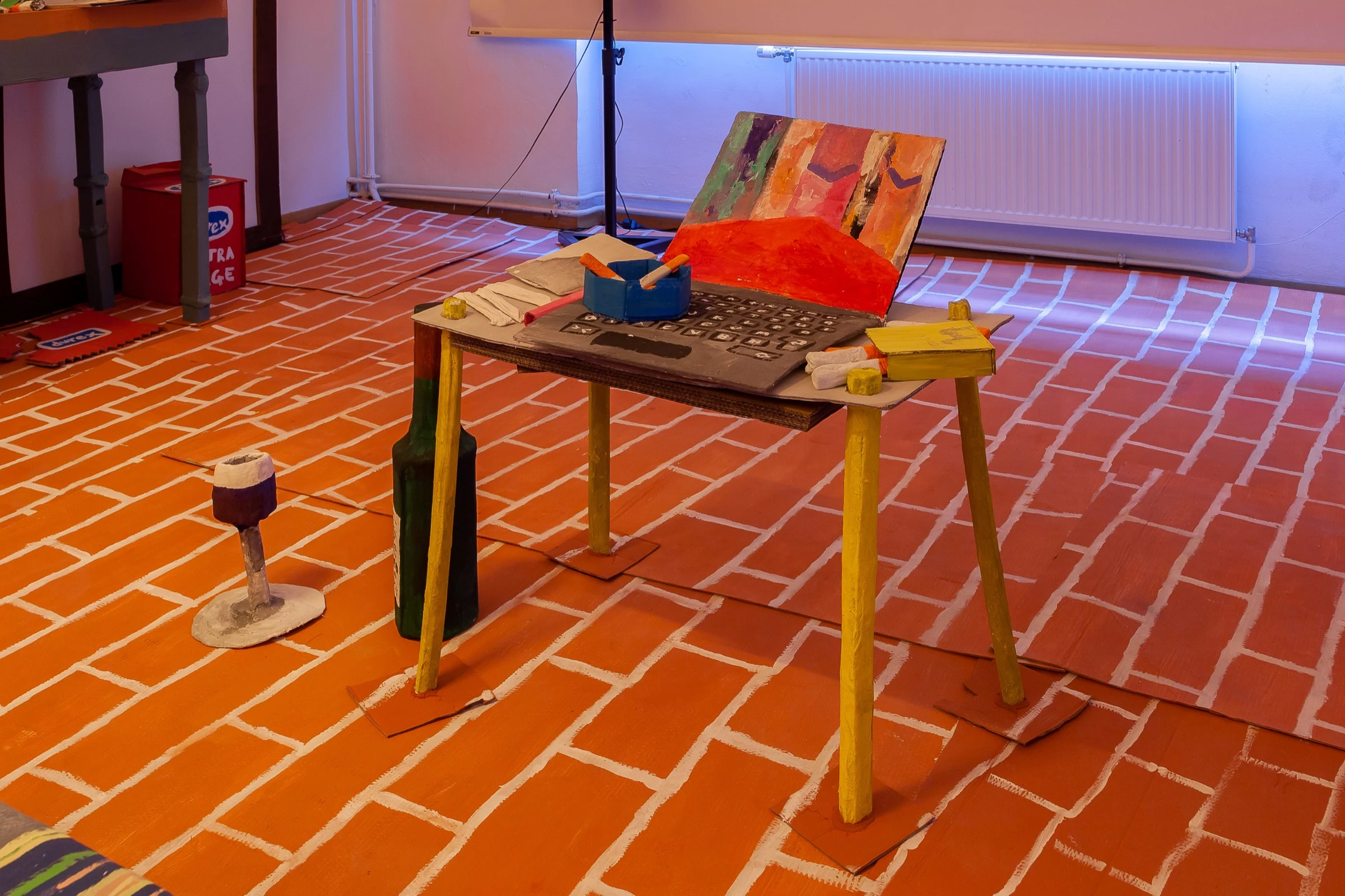 A cardboard laptop and ashtray on a handmade table in the installation.