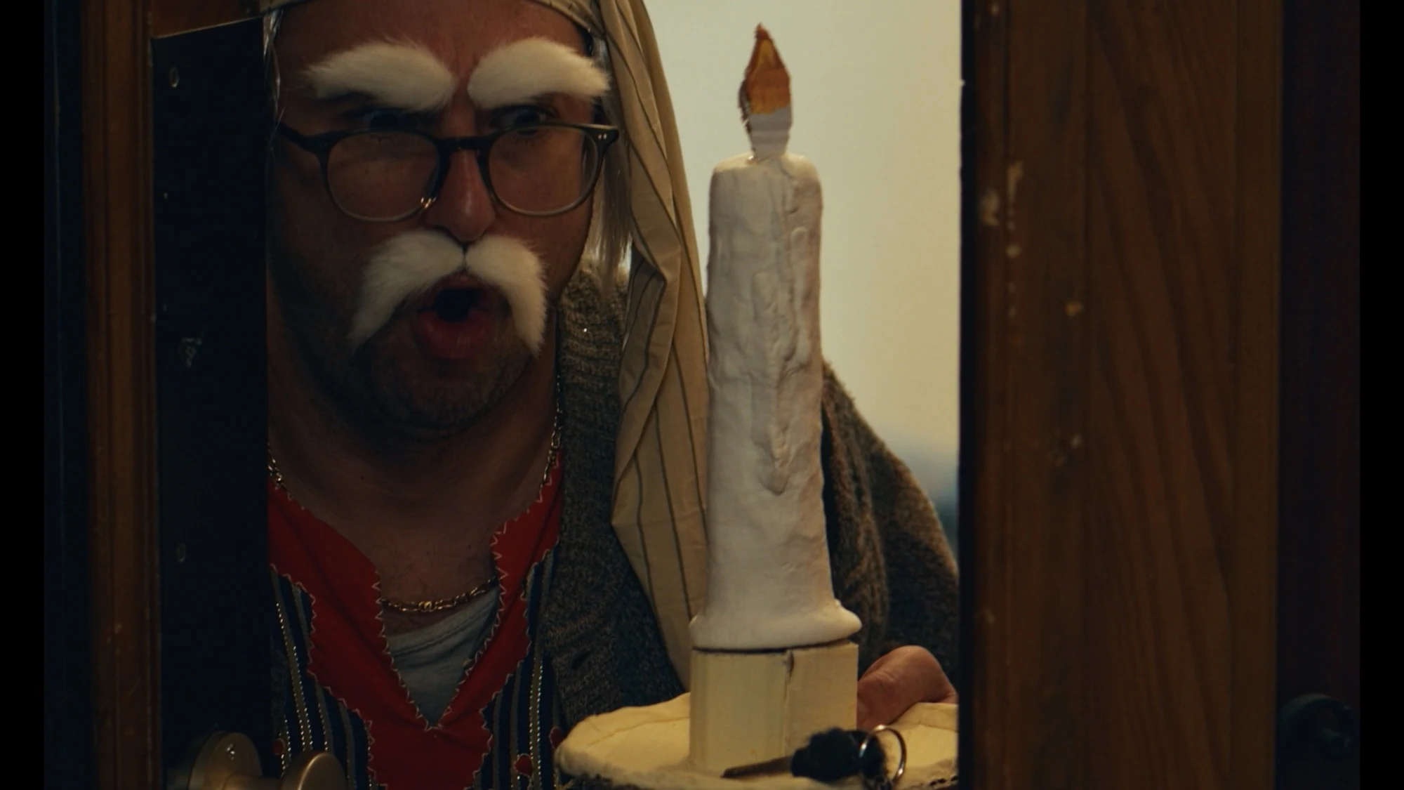 A costumed character with white eyebrows and a moustache holding a handmade candle in a doorway.