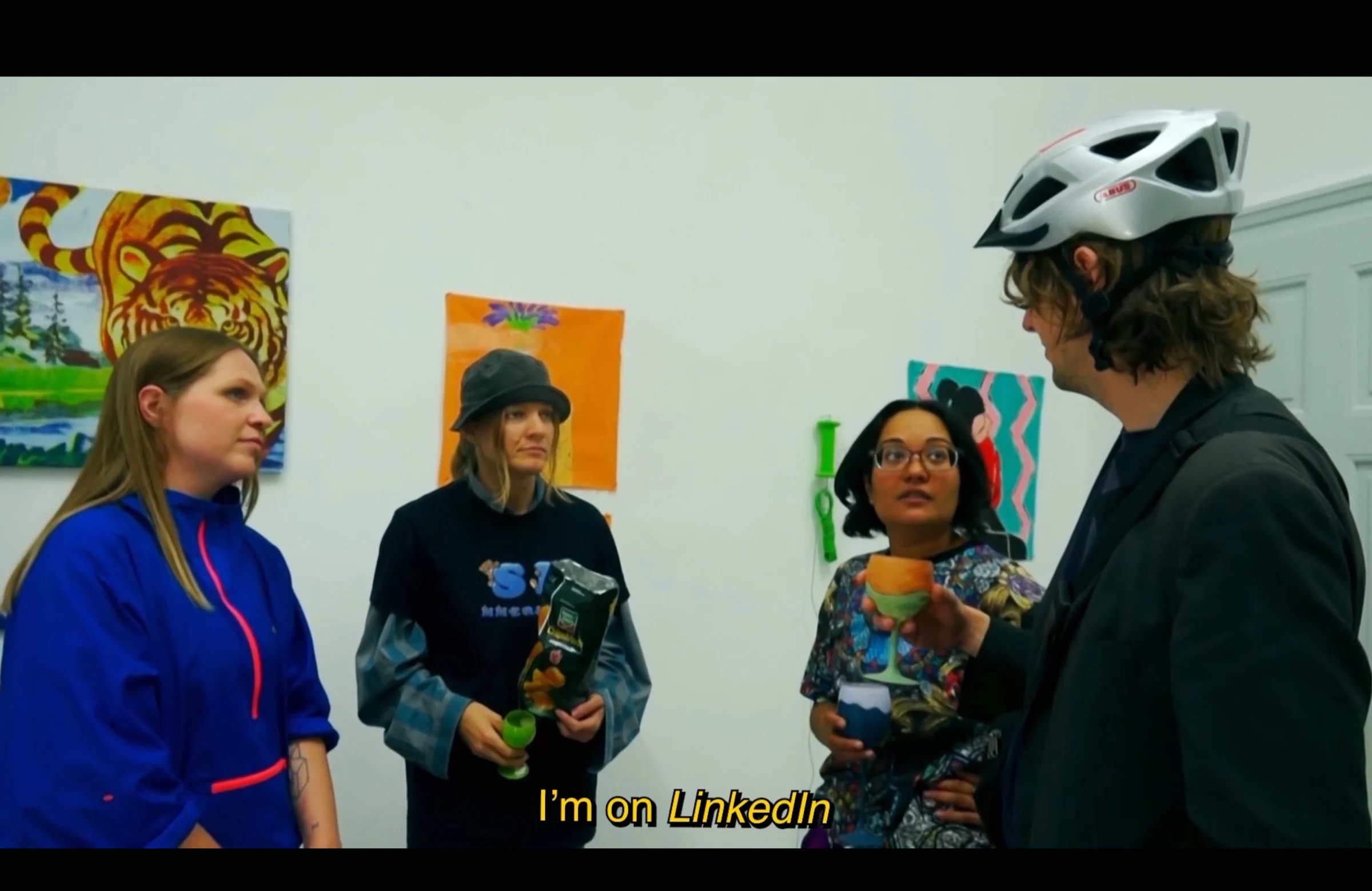 Four characters talking at an art exhibition, one wearing a bicycle helmet and holding a colourful glass.