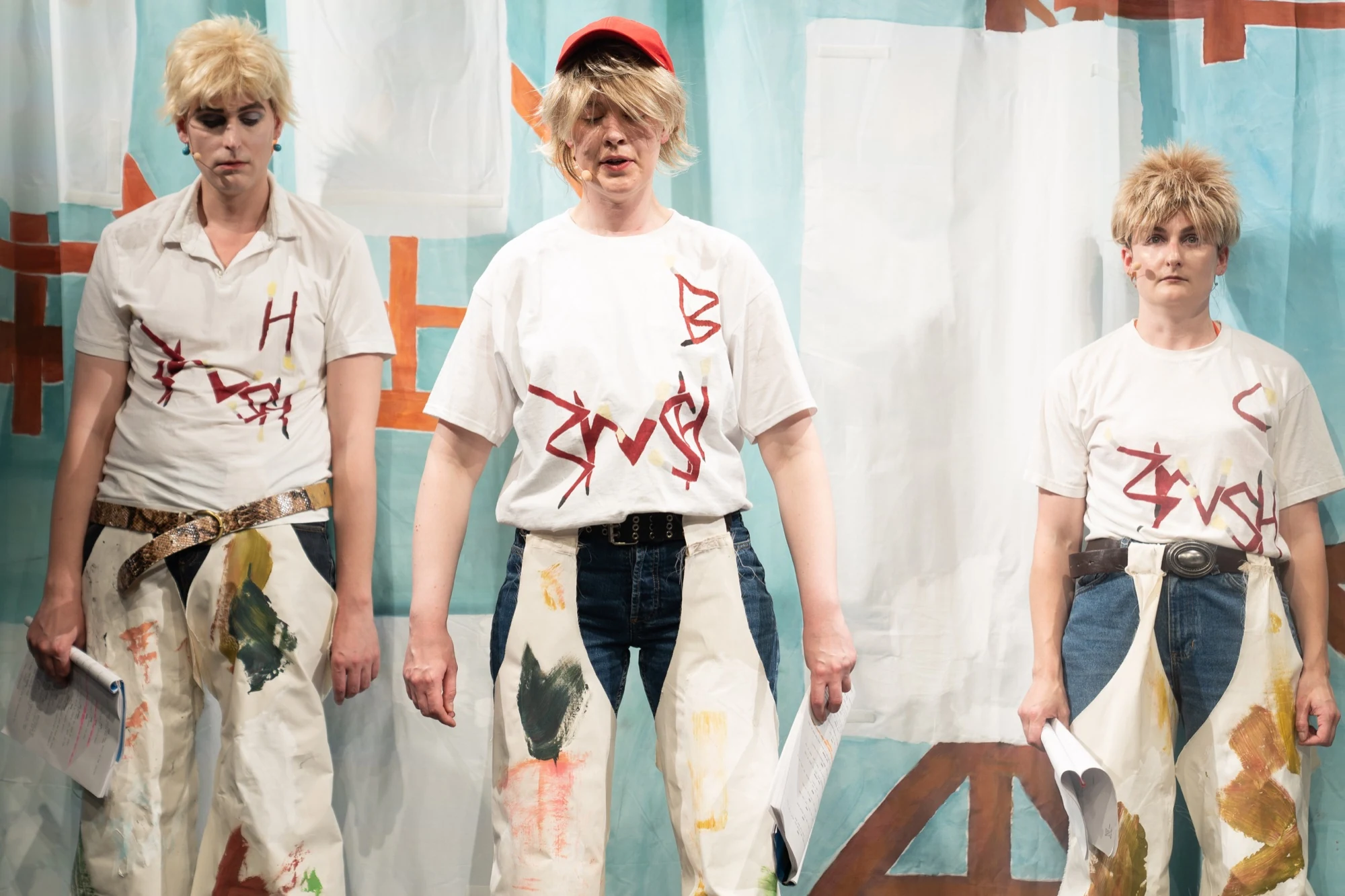Three performers in blond wigs and painted aprons holding scripts.