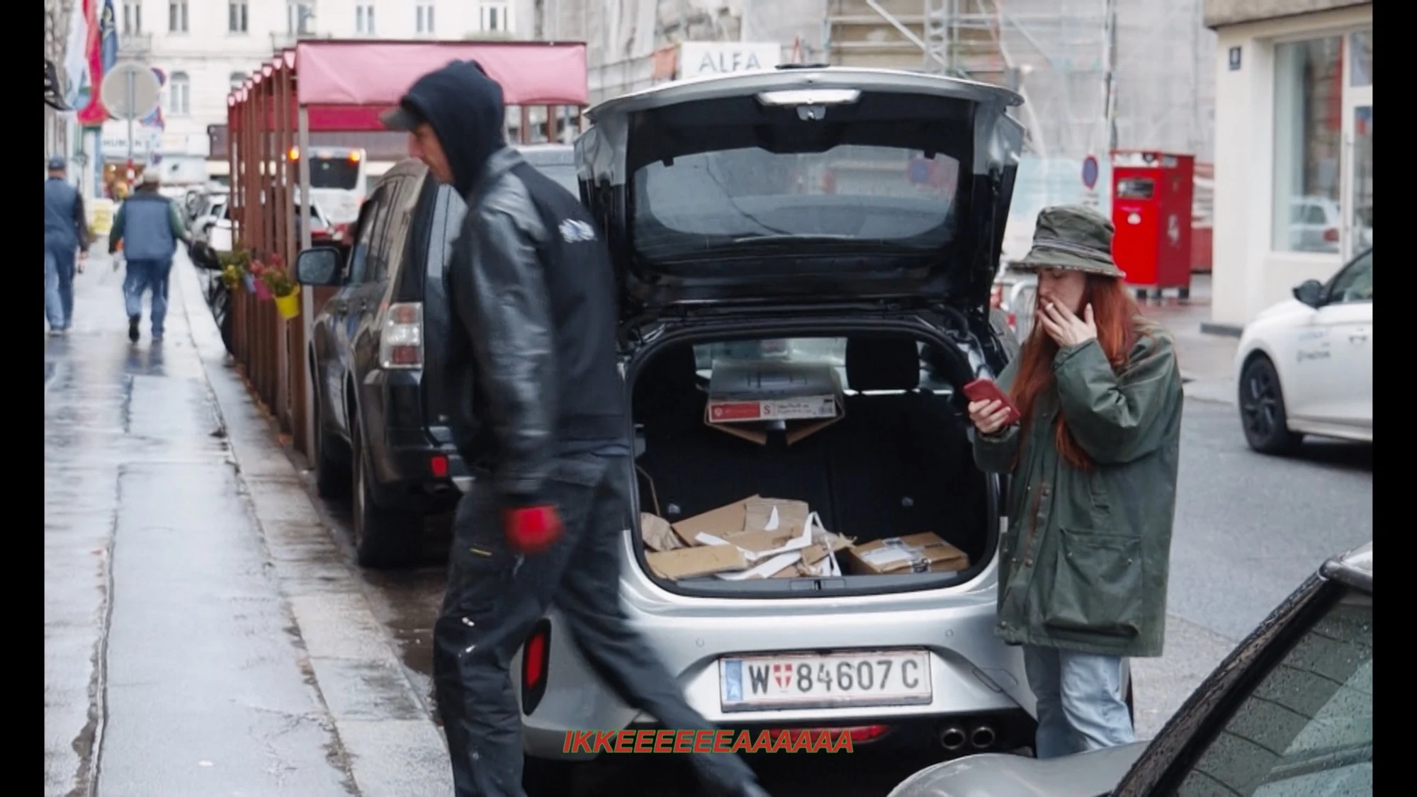 Charlotte holding a phone beside an open car boot on a rainy street.