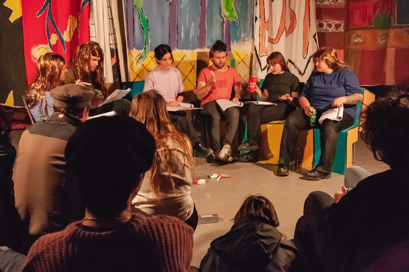 Performers seated on a colourful handmade couch with scripts, watched by an audience.