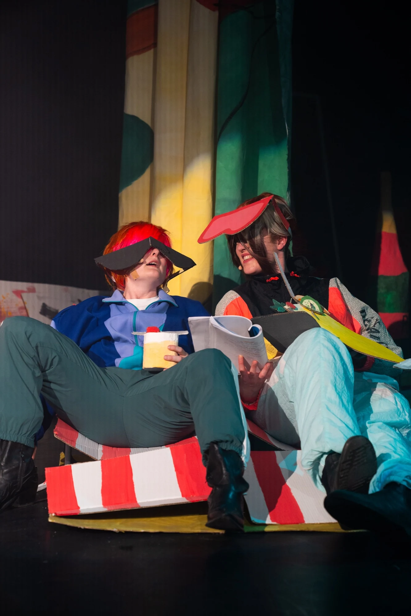 Two seated performers wearing oversized cardboard sunglasses and holding scripts.