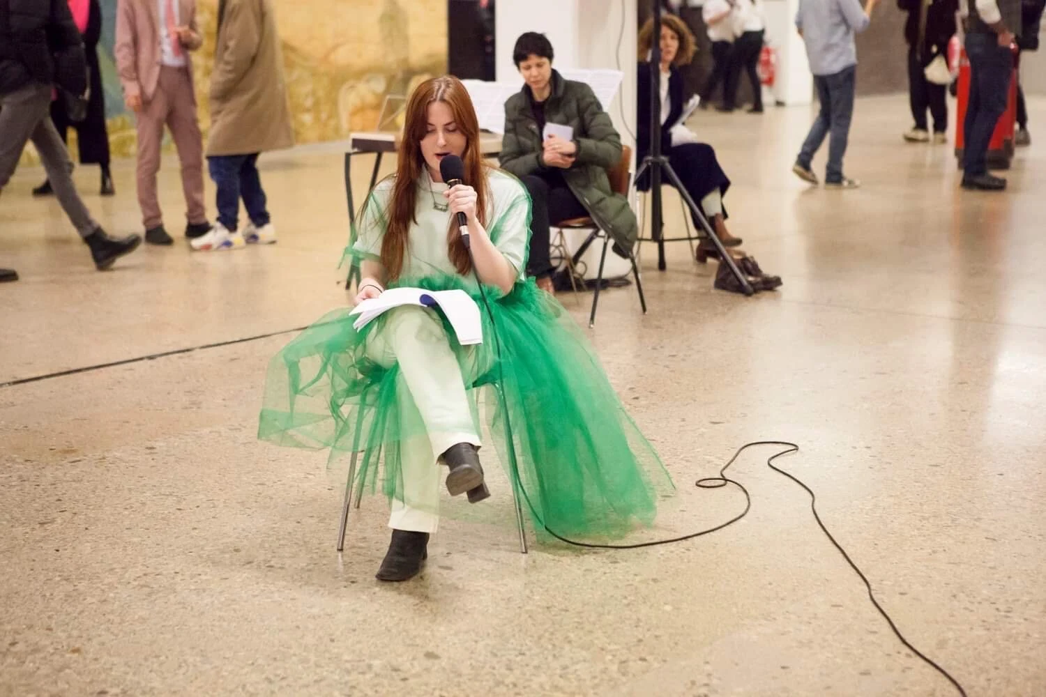 Charlotte Gash seated in a green tulle costume and reading into a microphone before a gallery audience.