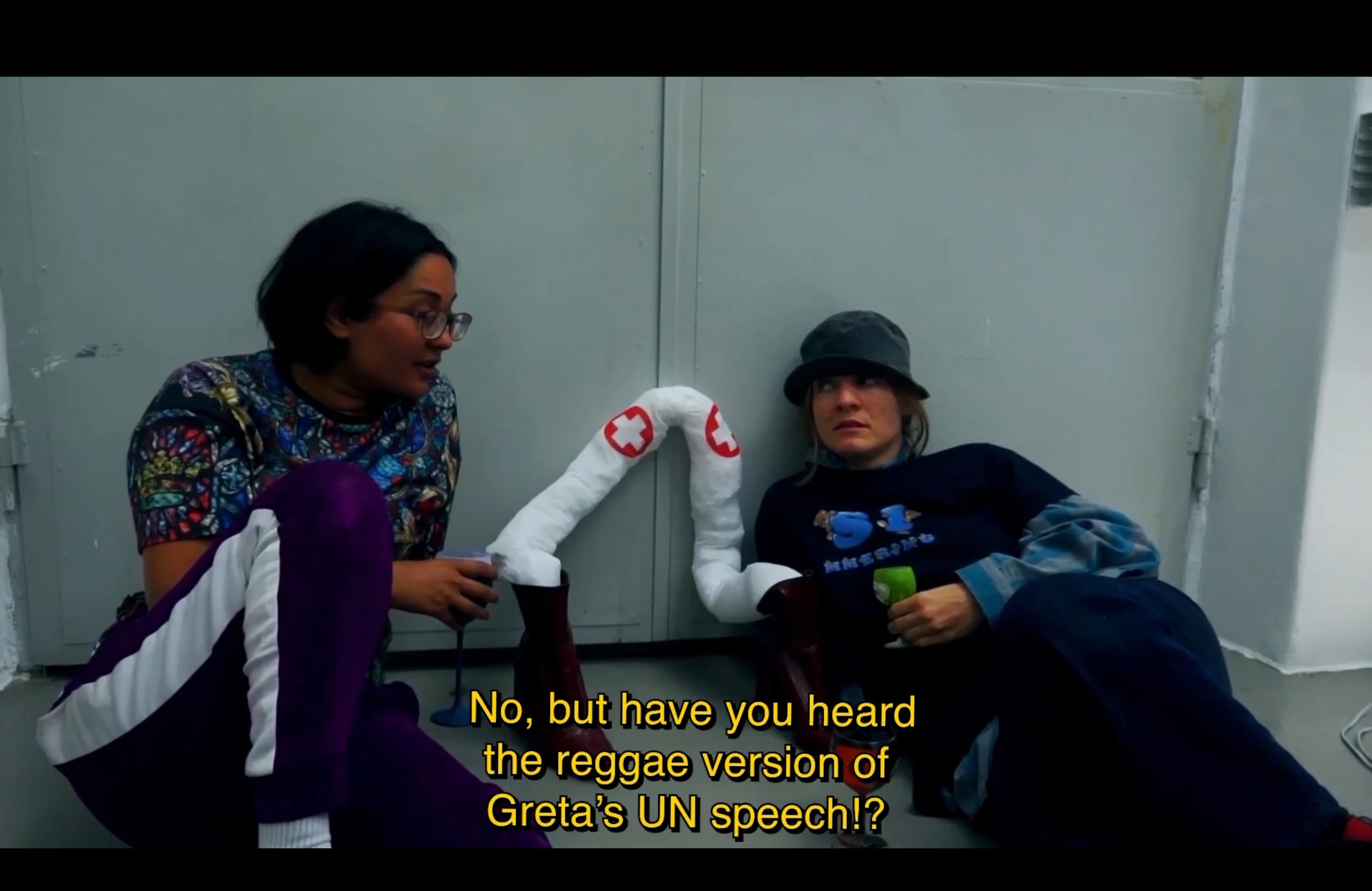 Two characters seated on the floor beside sculptural white arms, discussing a reggae version of Greta’s UN speech.