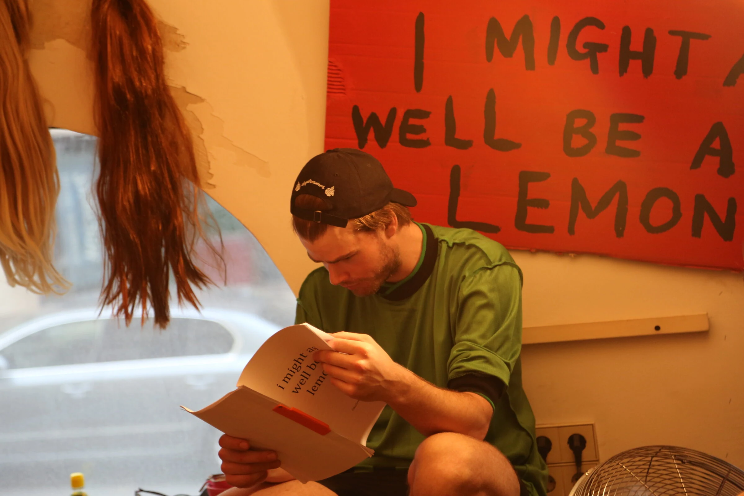 A performer in a green shirt and black cap reading the script beneath the red title sign.