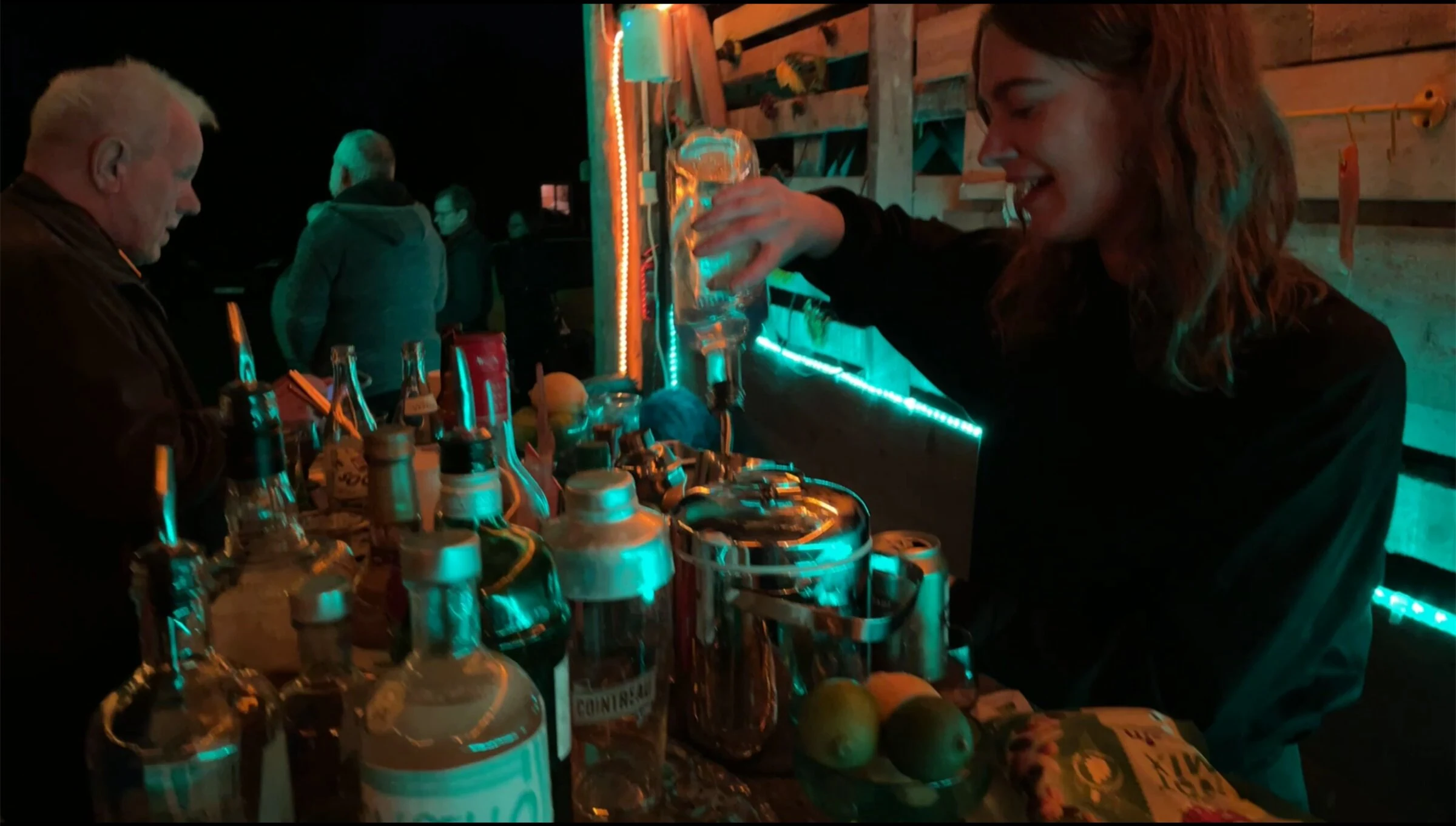 A person pouring a drink at a bar lit by turquoise strip lights.