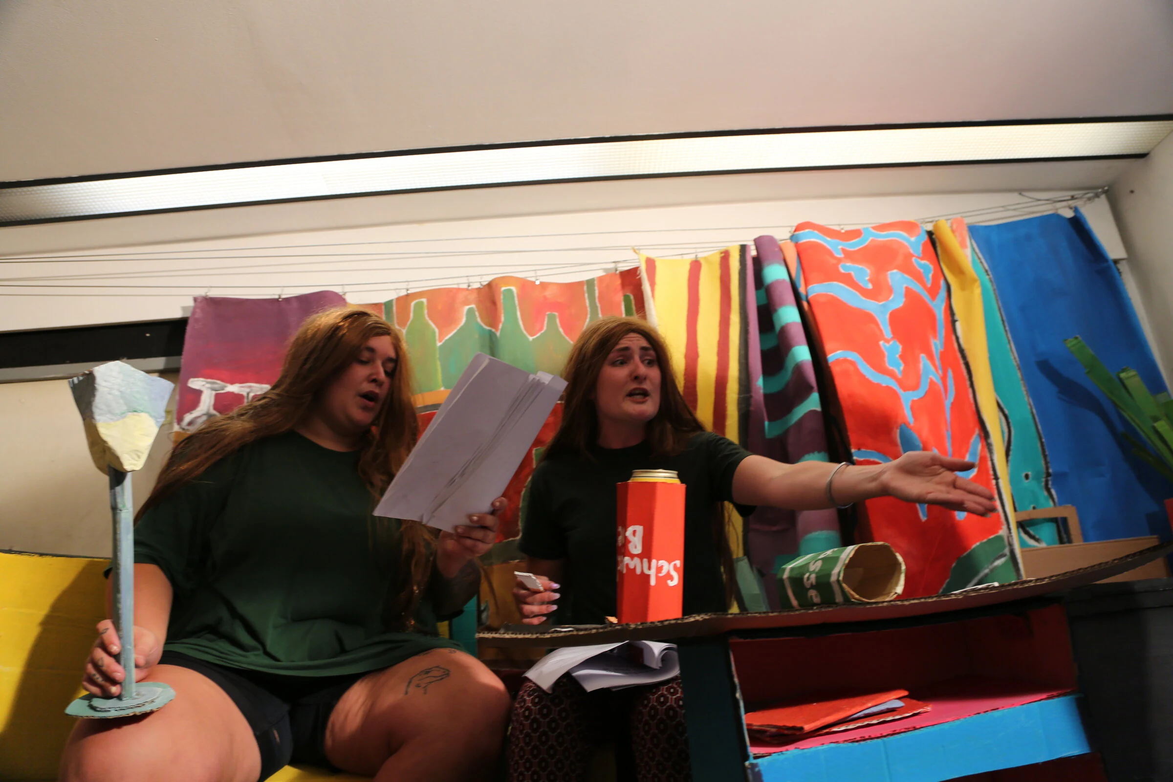Two seated performers with scripts and a cardboard cocktail glass in front of colourful painted curtains.