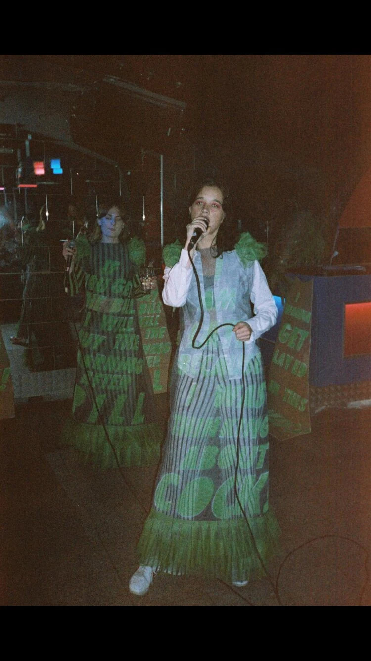 Two performers in dresses with projected green lettering, performing with a microphone.