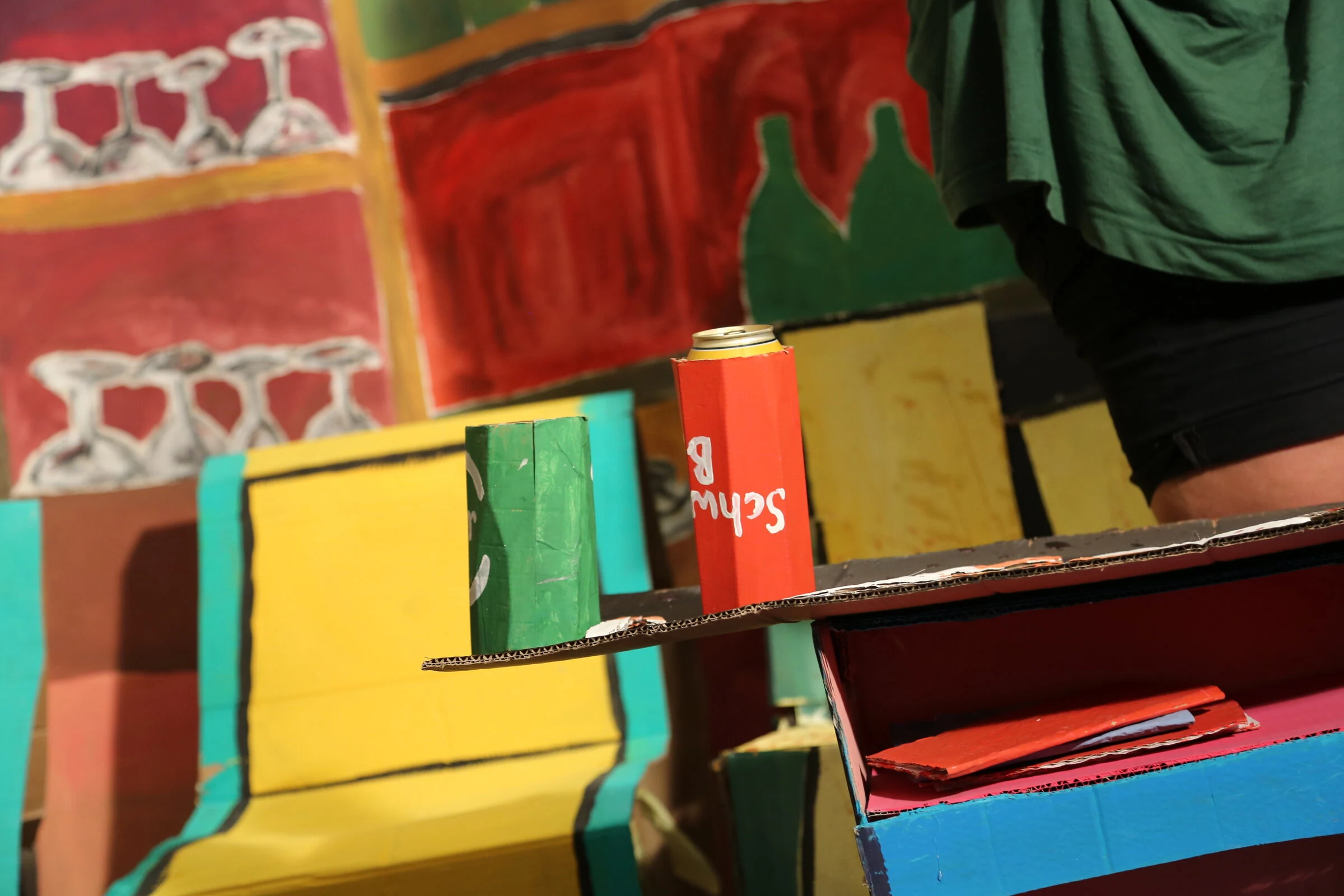 Red and green cardboard drink holders on a handmade table beside a yellow cardboard sofa.