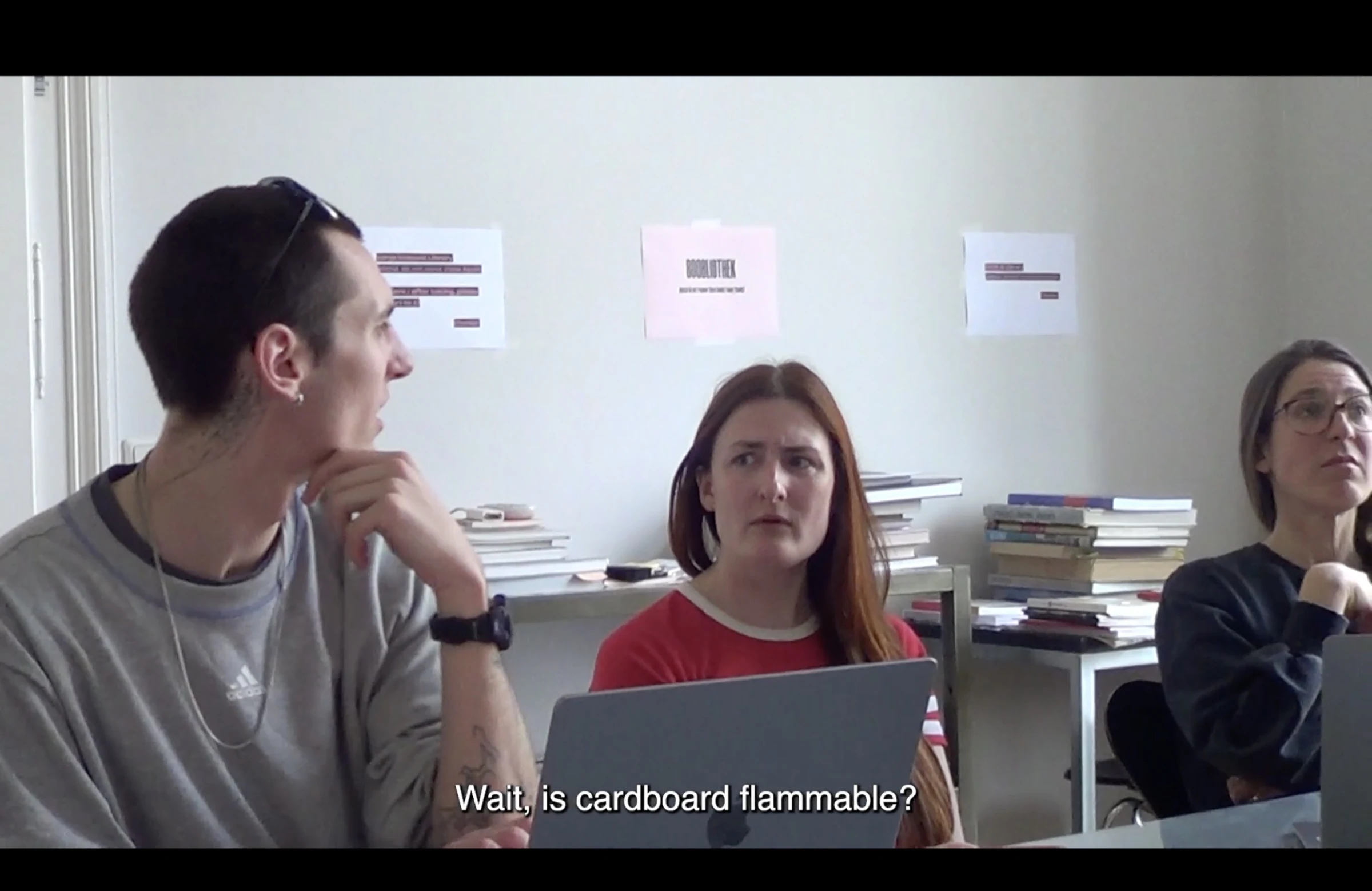 A film still of three people in a meeting with the subtitle Wait, is cardboard flammable?