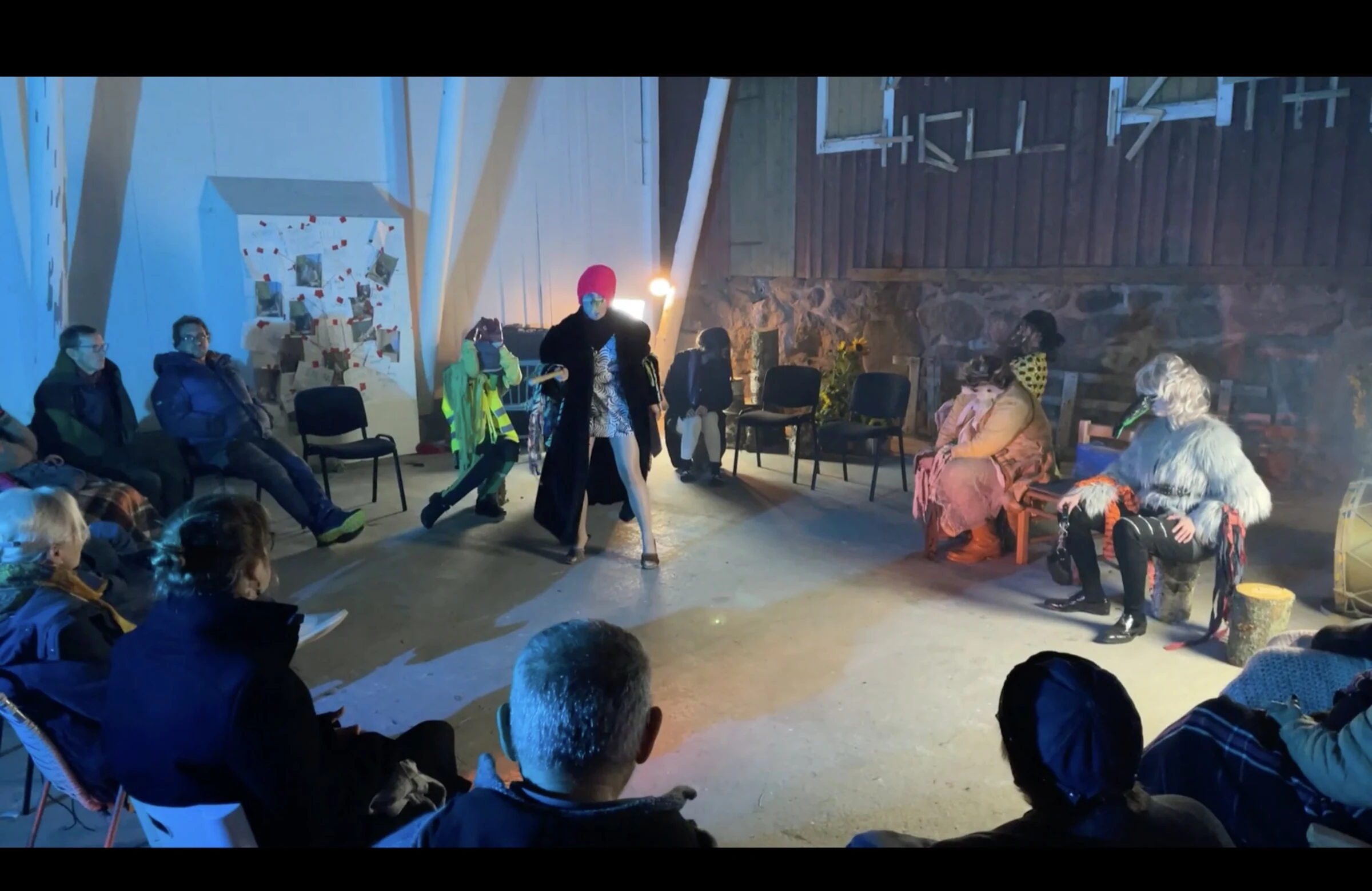 A performer standing with a staff amid the seated cast and audience.
