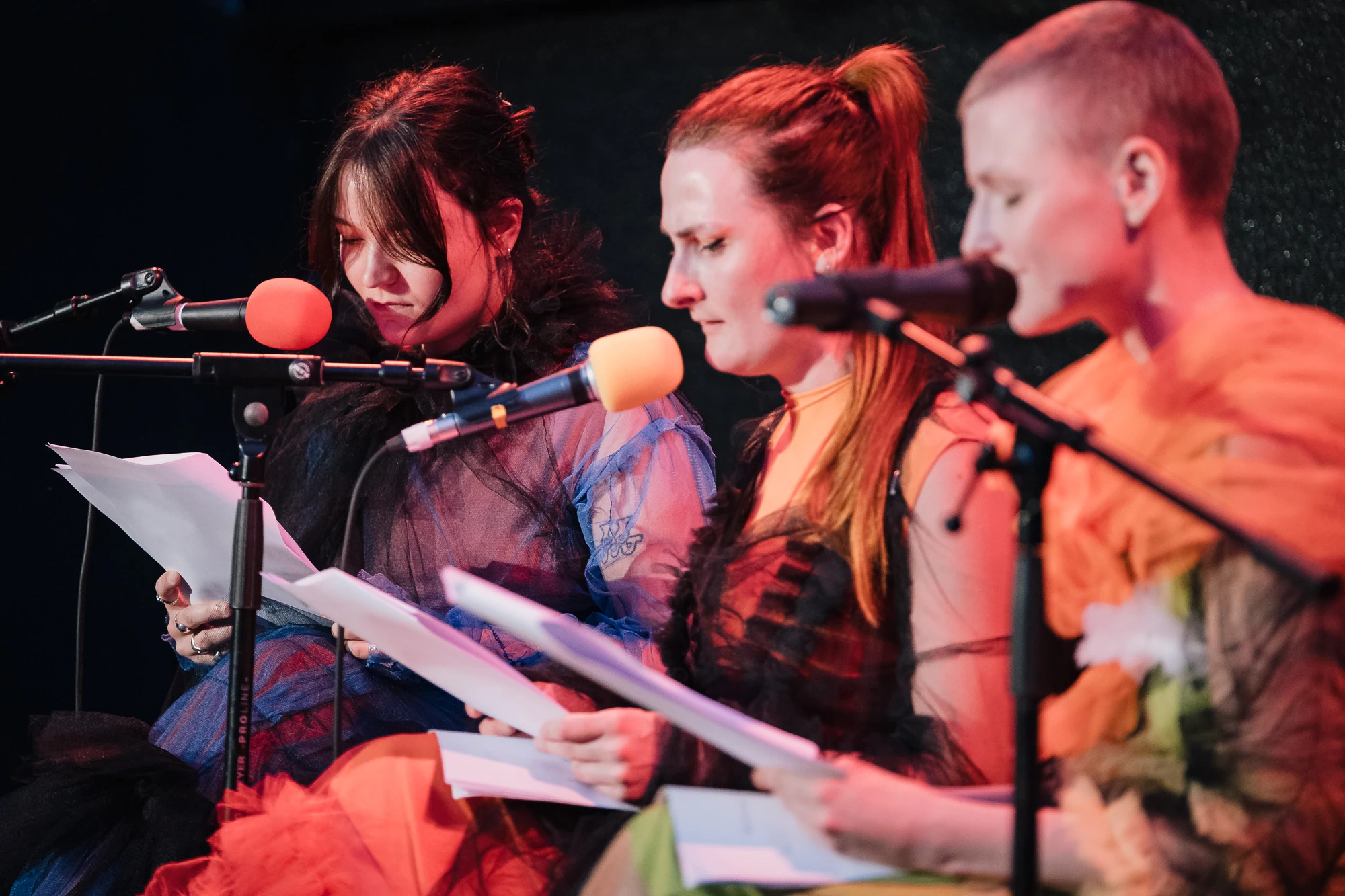 Three performers reading scripts into microphones under red stage lighting.