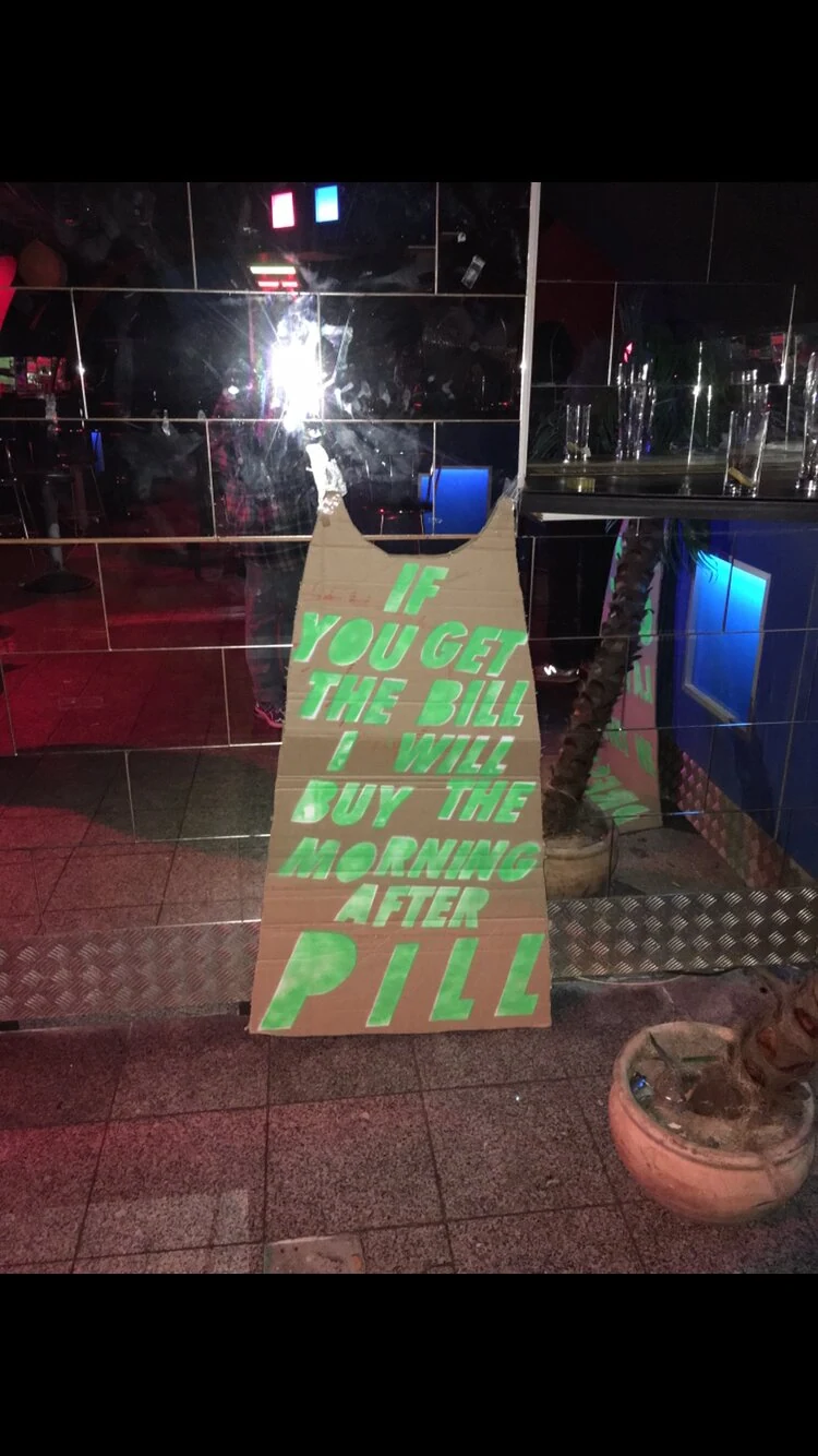 A cardboard dress with green lettering displayed against a mirrored bar wall.