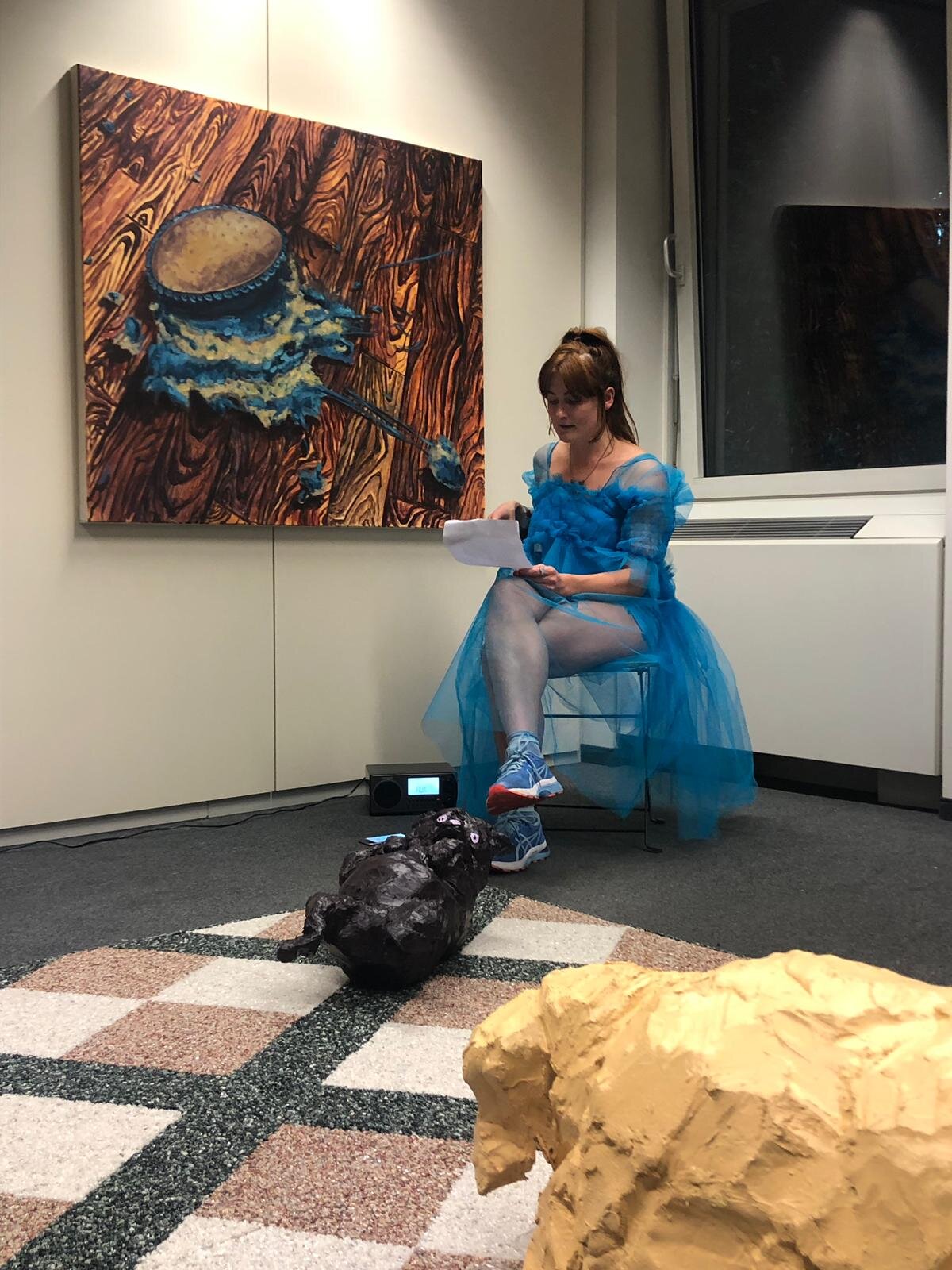 A performer in a blue tulle costume reading beside a painting and sculptures in a gallery.
