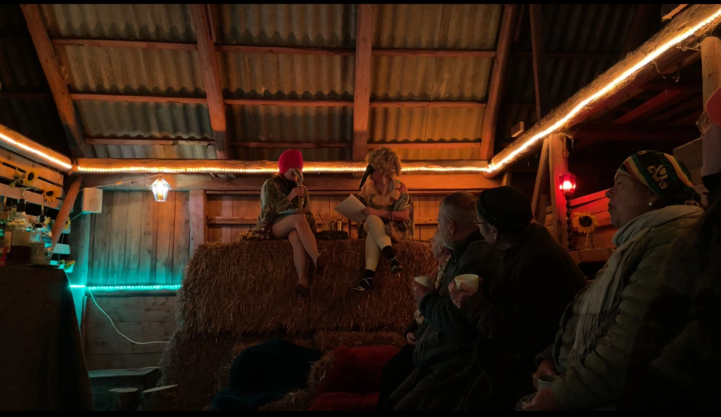 Two masked performers seated on straw bales beside audience members.