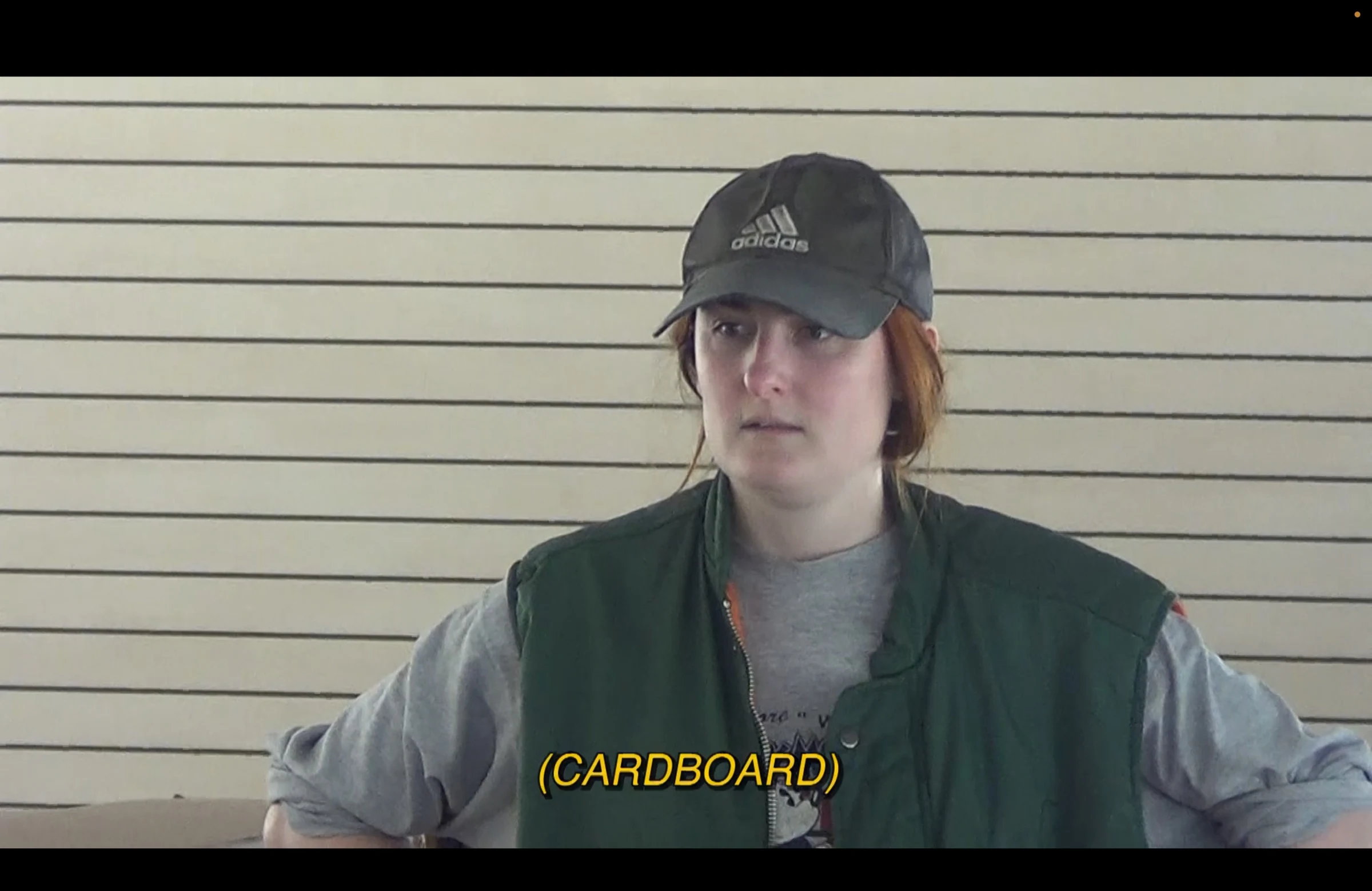 Charlotte wearing a cap and green vest in a film still with a cardboard subtitle.