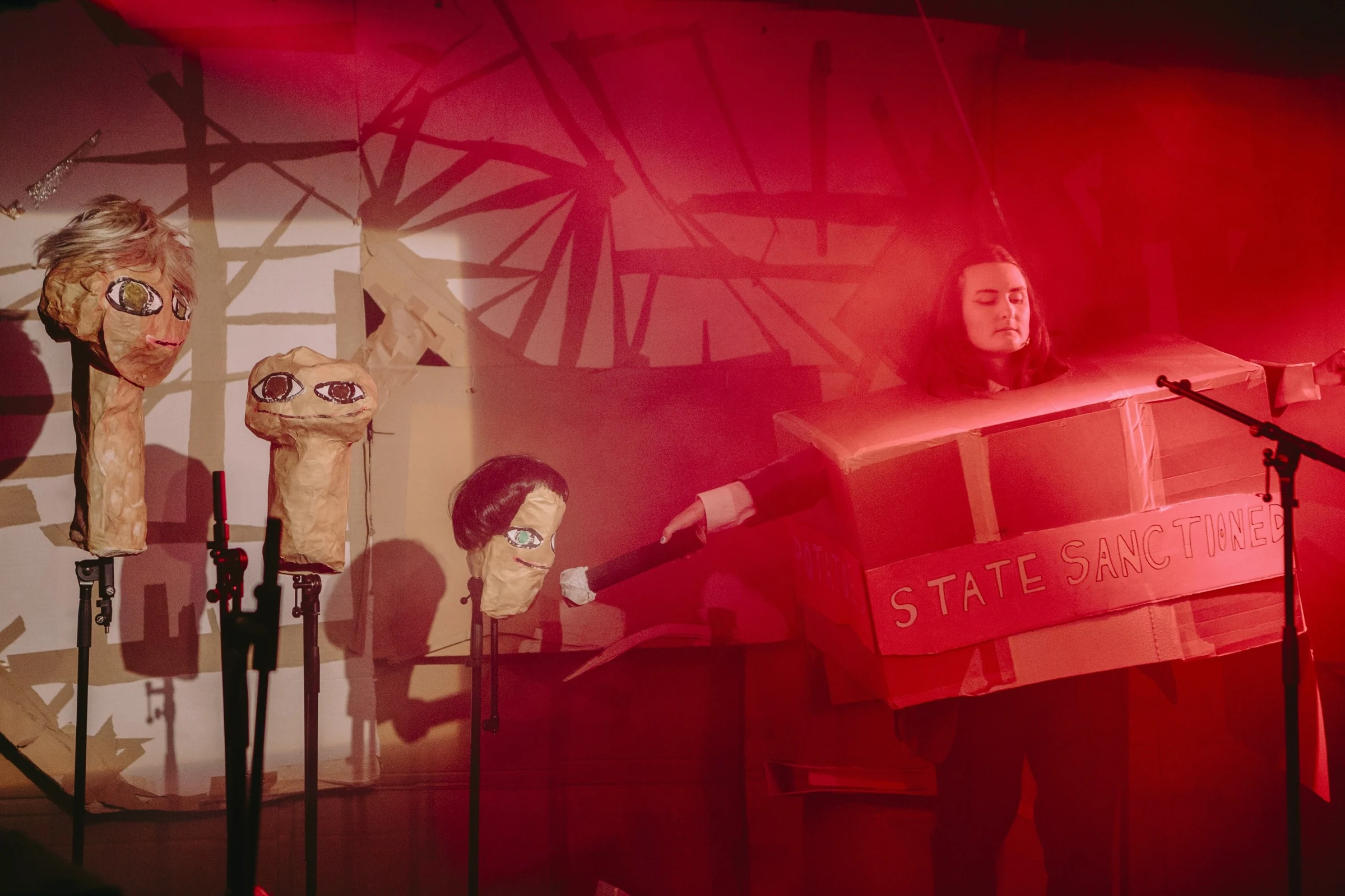A Gash Club performer in a cardboard costume beside handmade heads under red stage lighting.