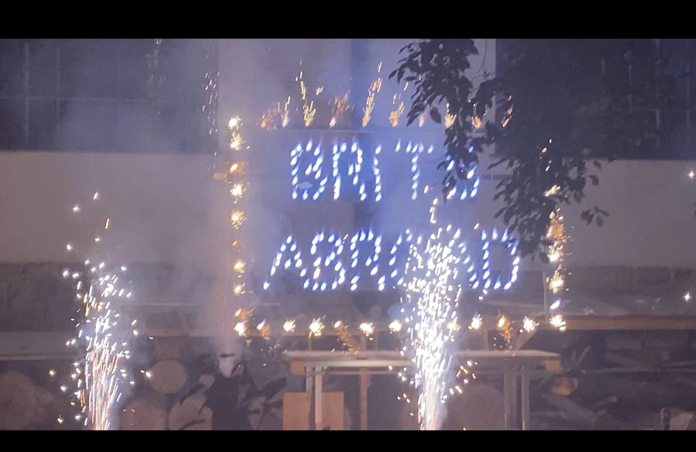 Sparks bursting around the illuminated BRITS ABROAD sign outside at night.