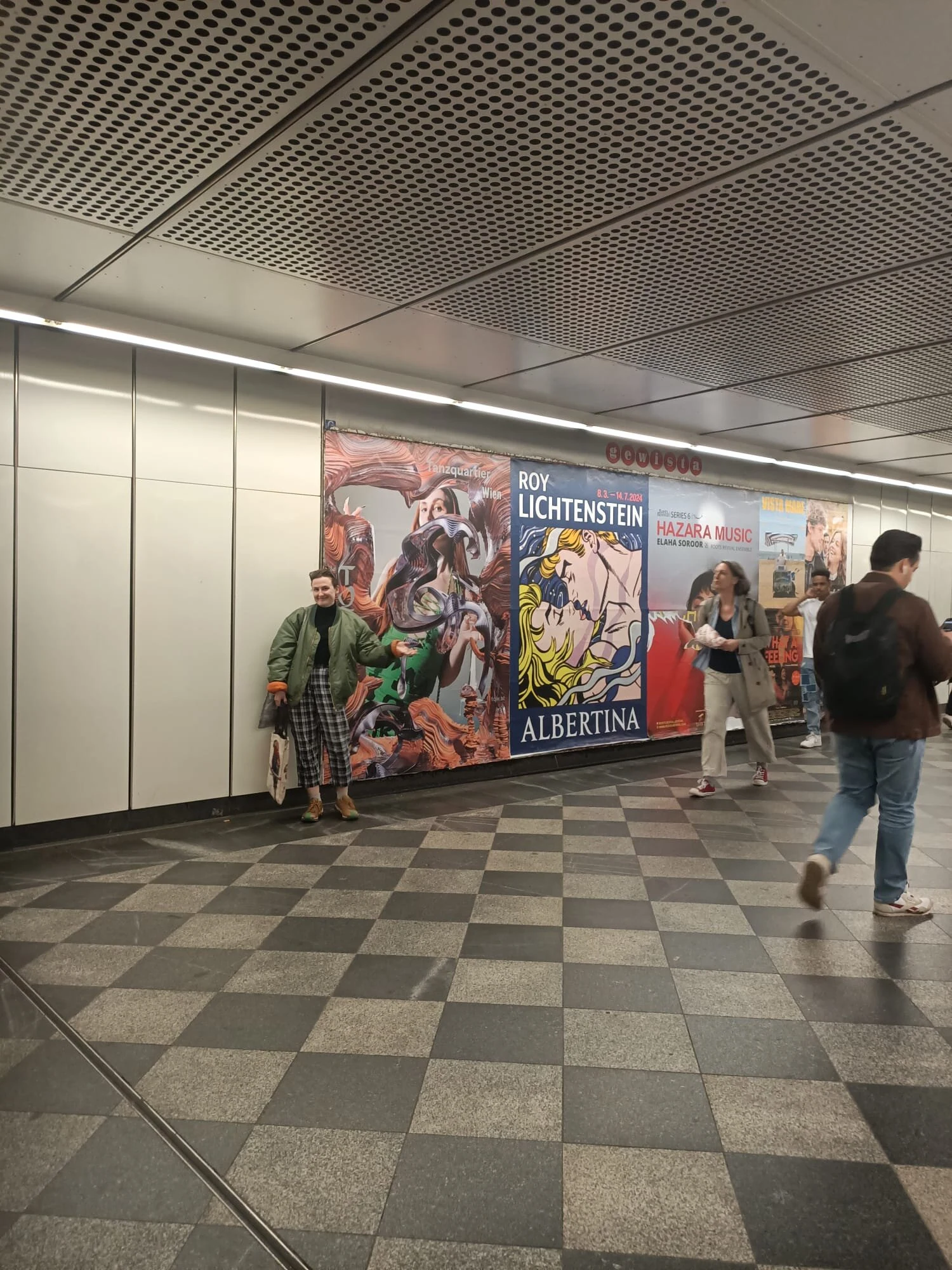 A person standing beside the green Tanzquartier poster among advertisements in a station passage.