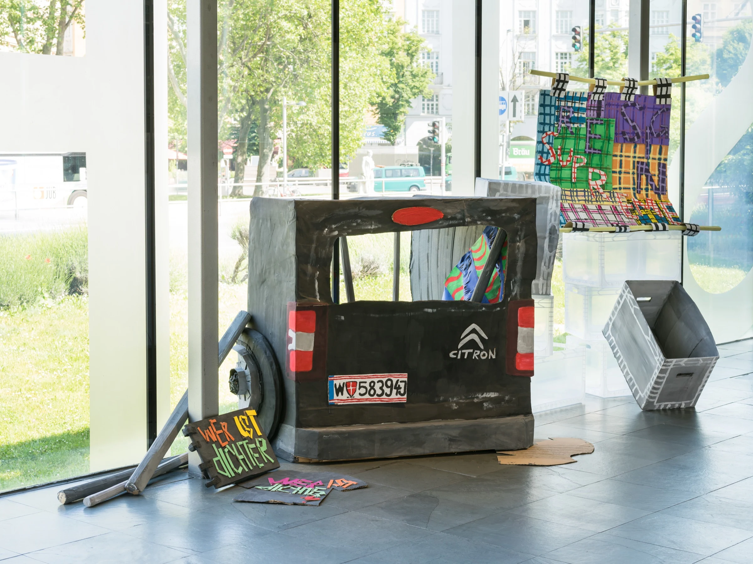 The rear of the cardboard van with colourful textiles, signs and a grey crate.