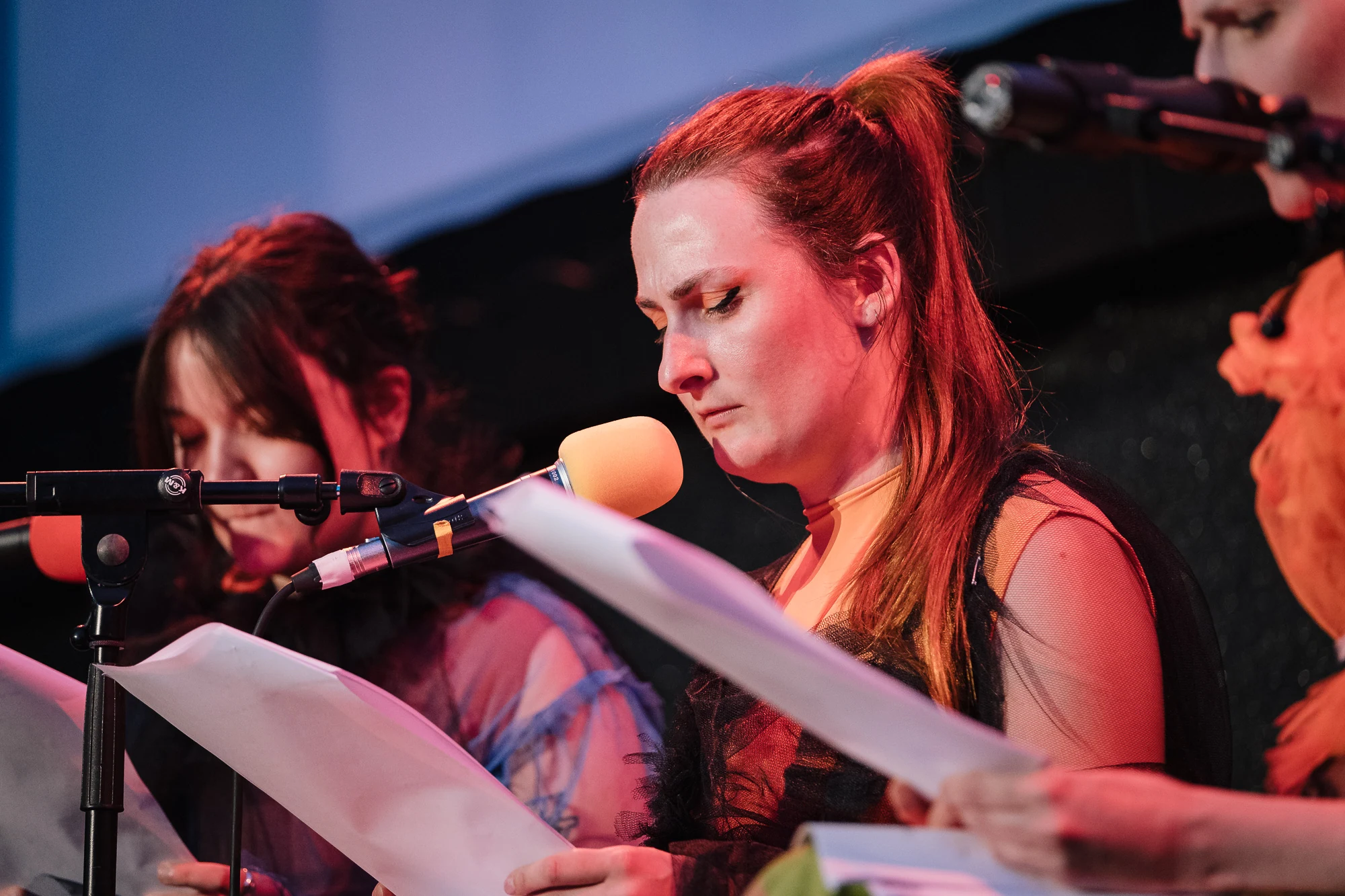 A close-up of a red-haired performer reading into an orange microphone on stage.