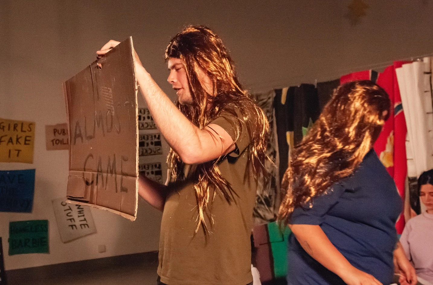 Performers wearing long wigs, with one holding a cardboard sign.