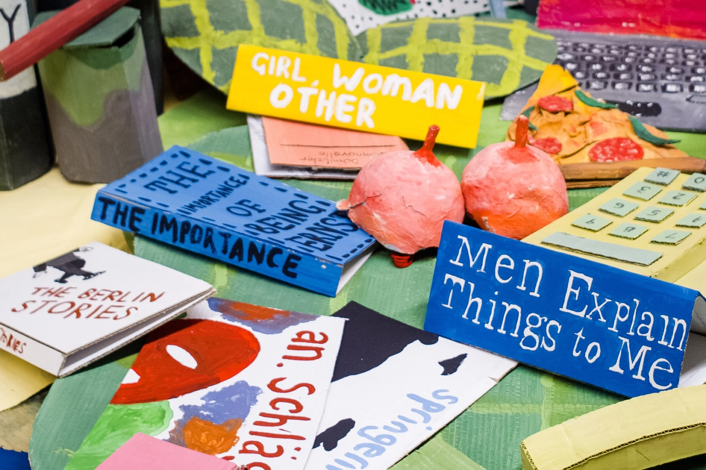 A close view of BEDROOM’s cardboard books, pink sculptures, pizza and telephone on a green table.