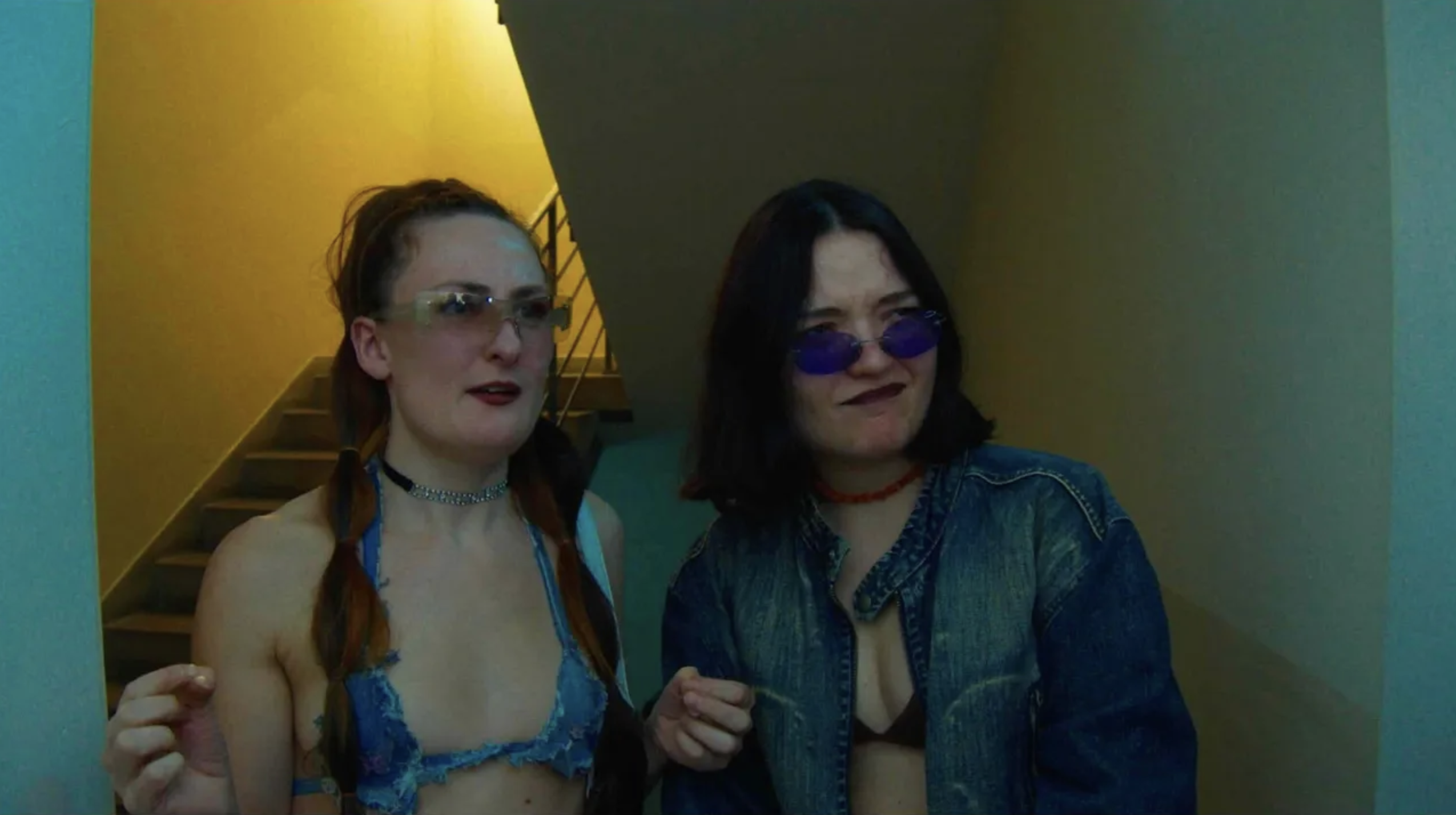 Two women wearing tinted glasses beside a yellow stairwell, one in a blue top and one in a denim jacket.