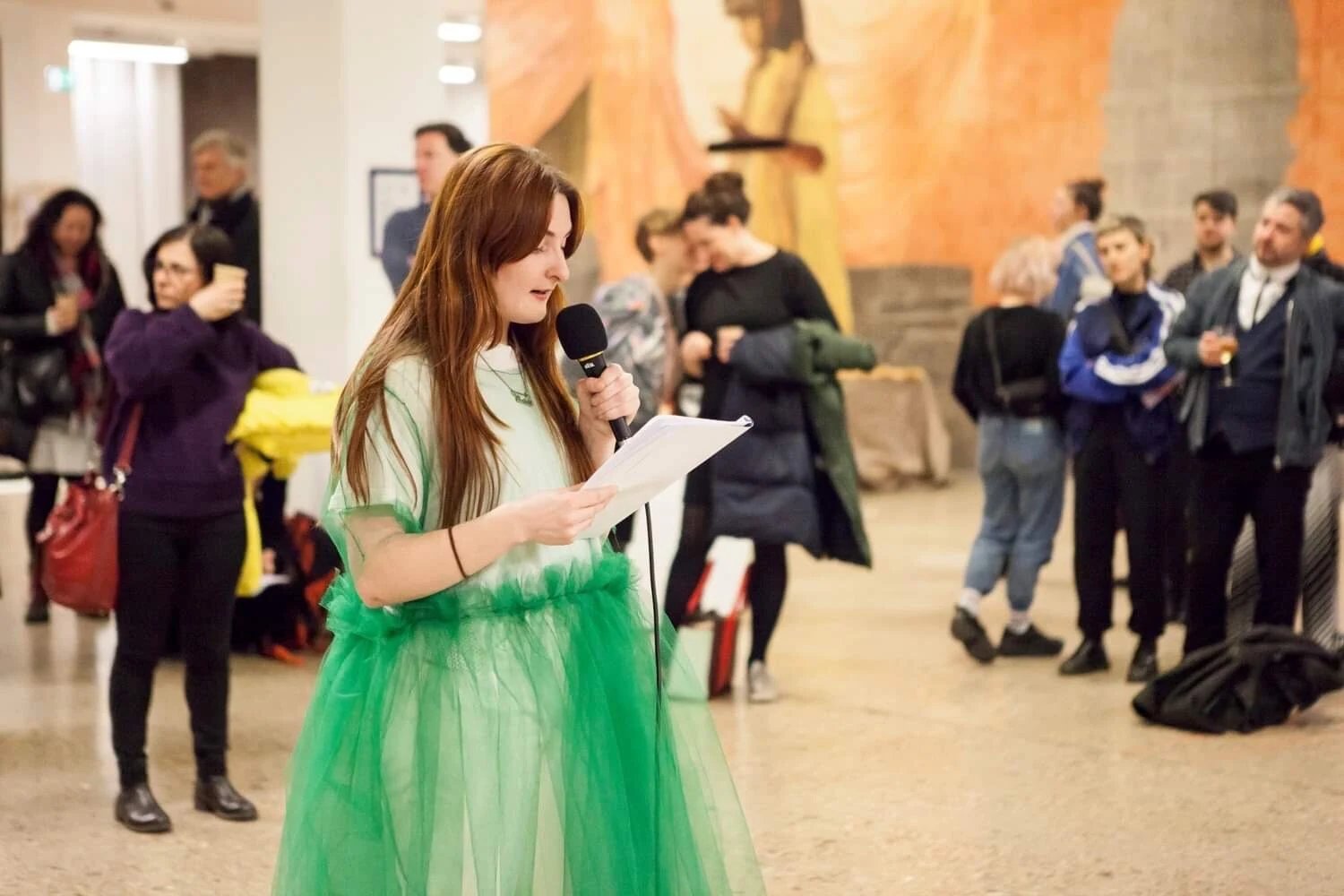 Charlotte Gash standing in a green tulle costume and reading into a microphone before a gallery audience.