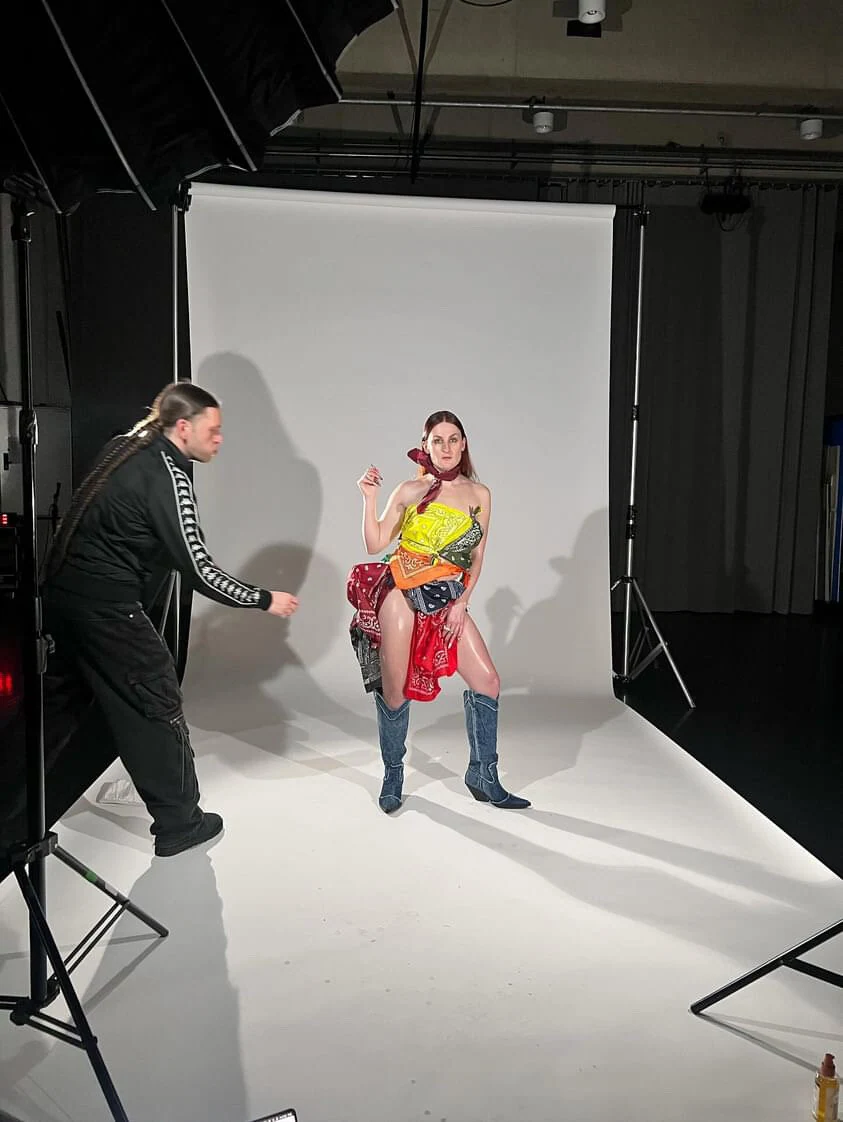 A wide studio view of Charlotte posing in bandanas and denim boots while a stylist adjusts the outfit.