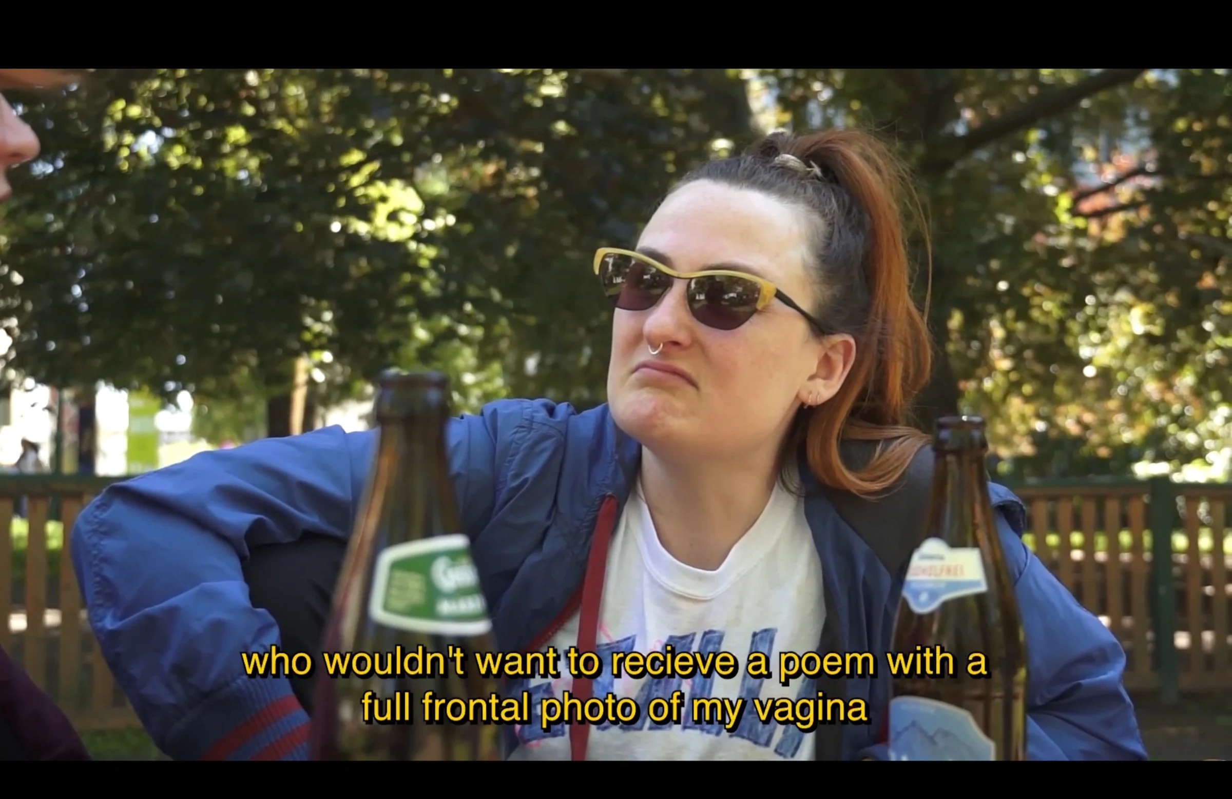 A character wearing yellow-framed sunglasses and a blue jacket sitting outdoors behind two beer bottles.