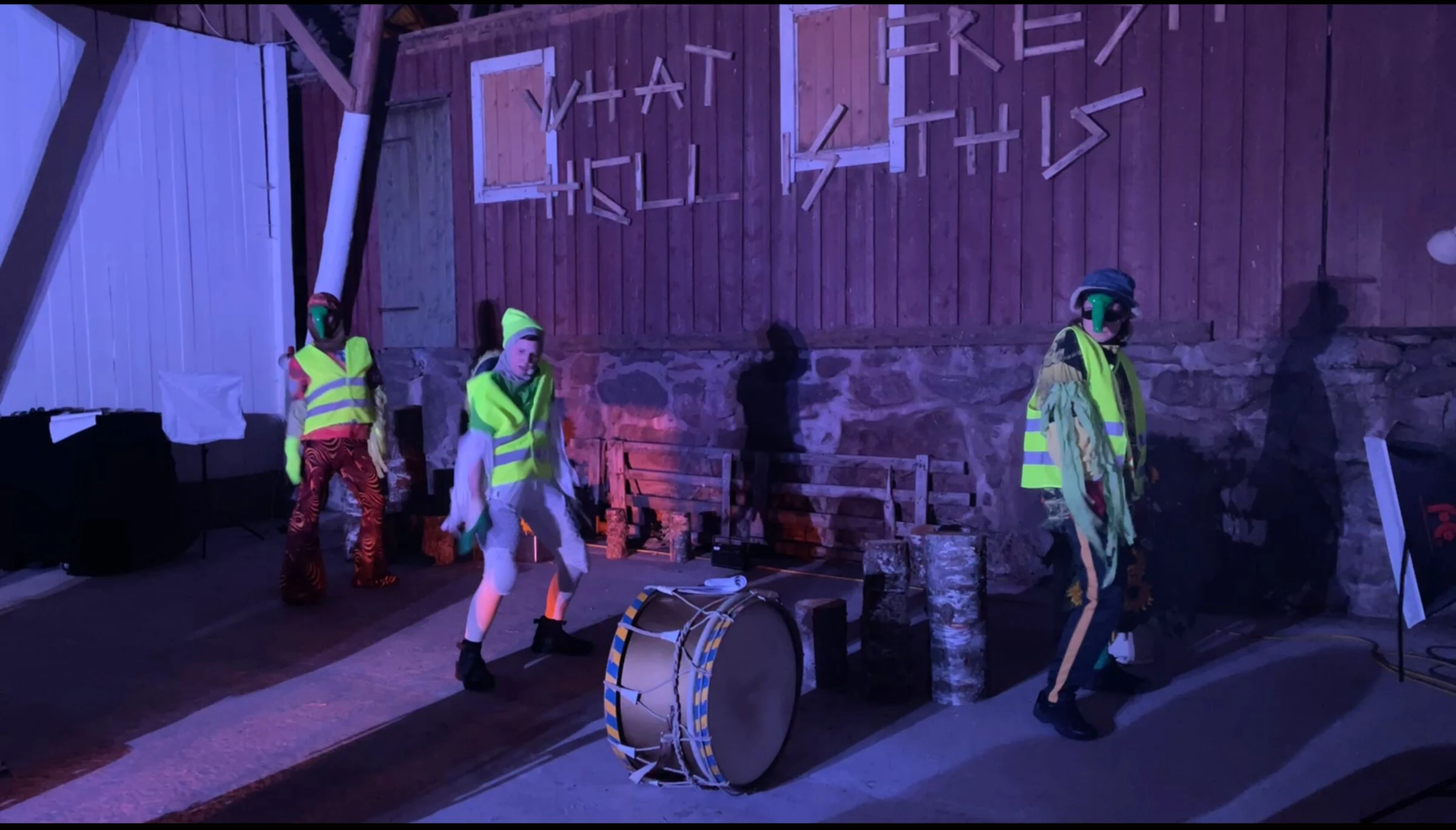 Three masked performers in fluorescent vests performing beside a drum under purple lighting.