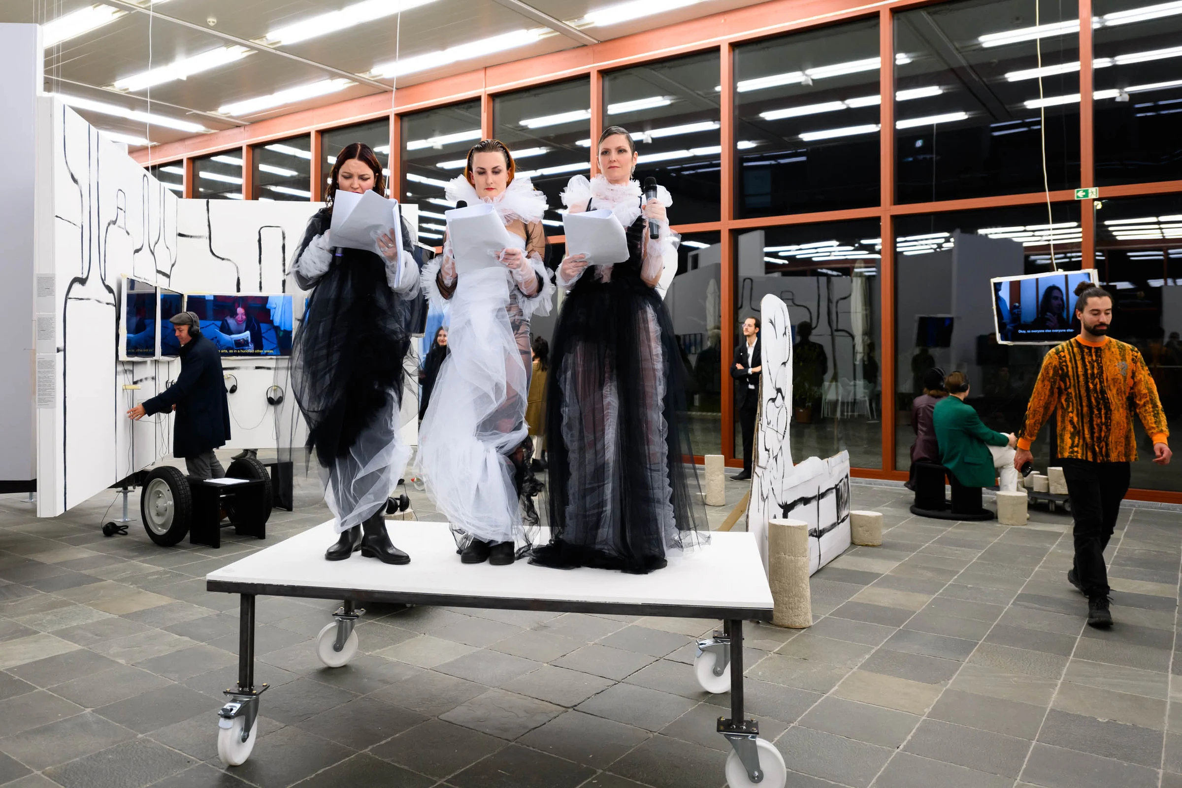 Three performers on a wheeled platform surrounded by video screens and cardboard sculptures.
