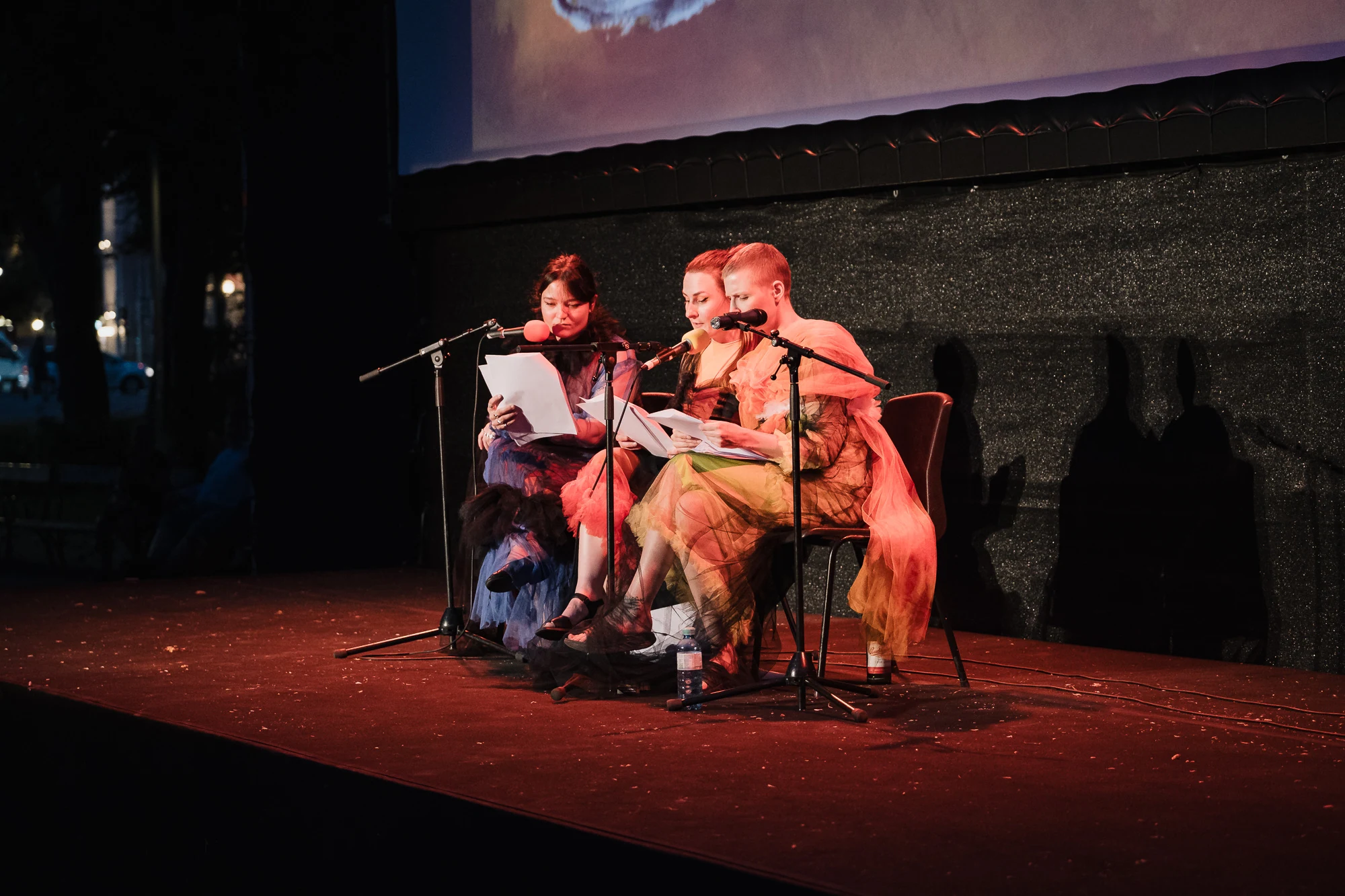 Three seated performers reading scripts on an outdoor stage at dusk.