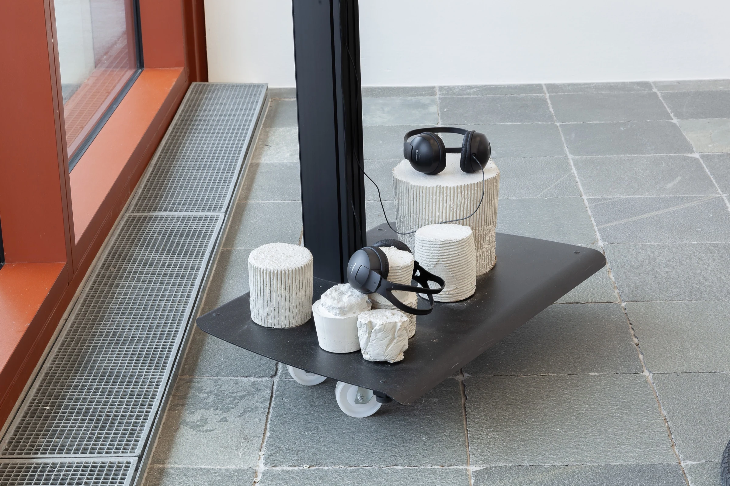 White cylindrical sculptures and headphones arranged on a wheeled platform beside the windows.