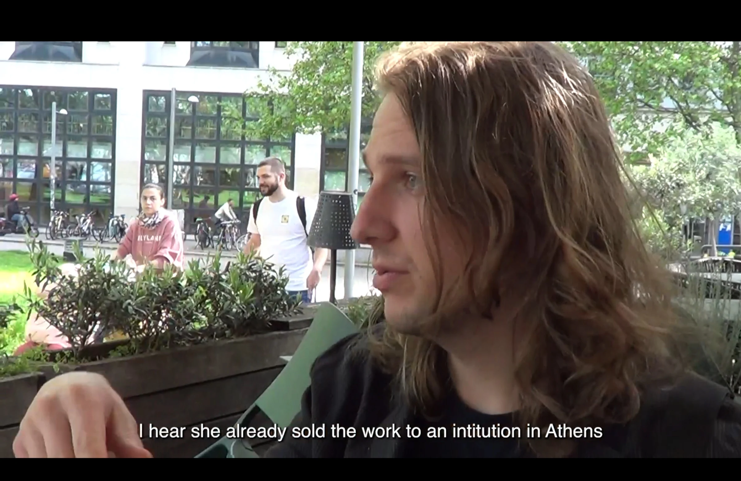 A long-haired character outdoors discussing the sale of a work to an institution in Athens.