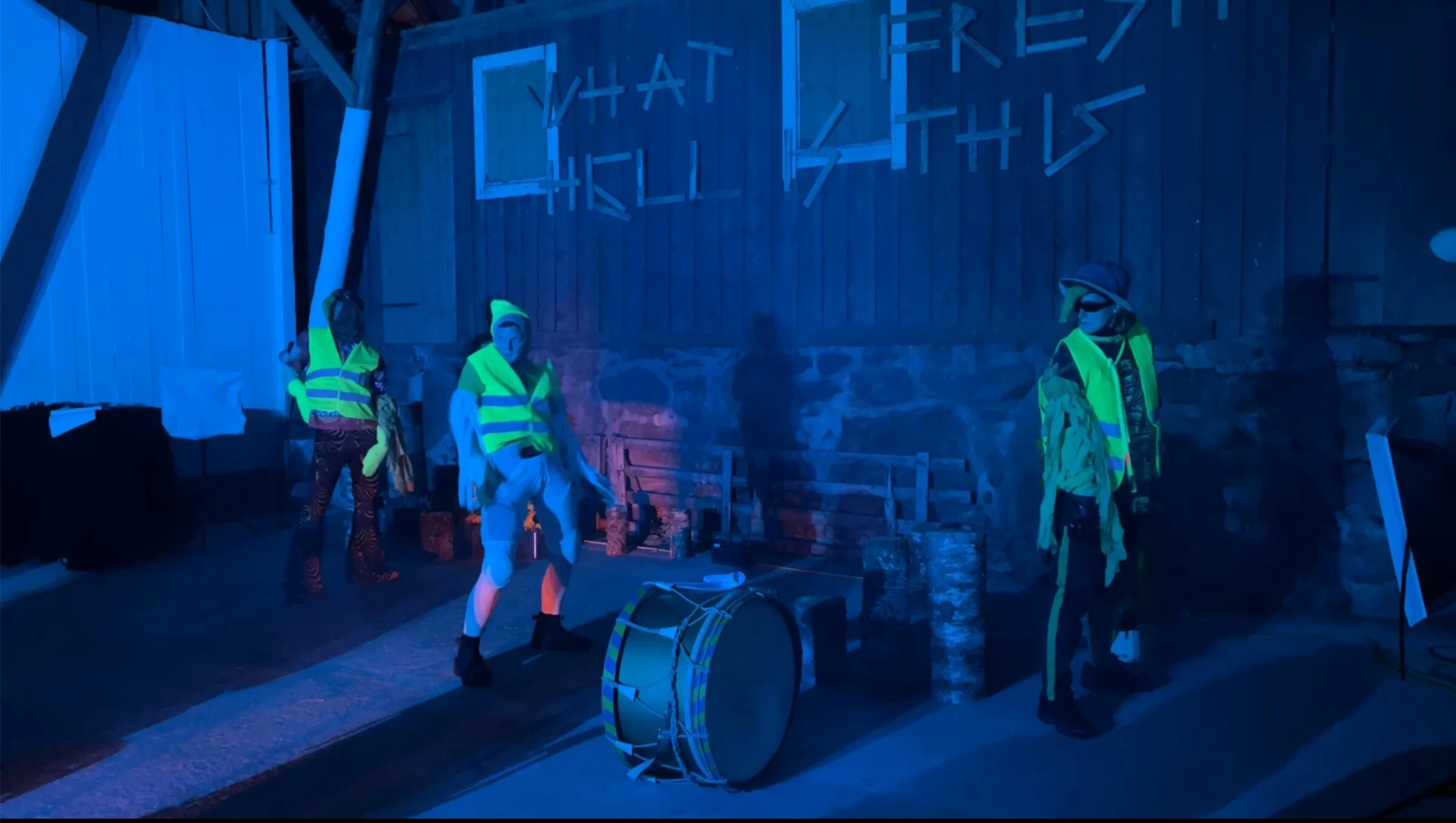 Three performers in fluorescent vests beside a drum under blue lighting.