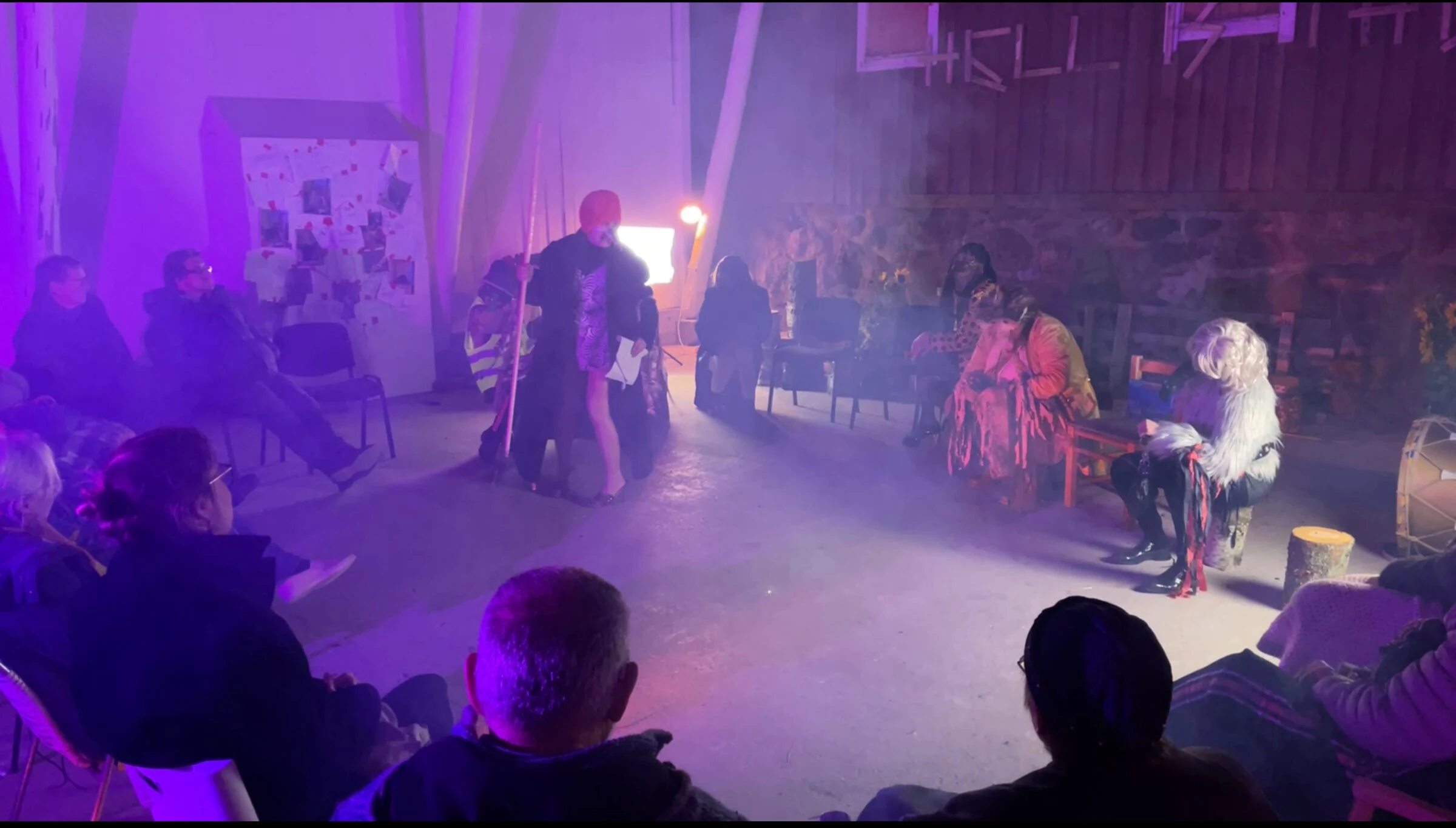 A masked performer holding a staff amid a seated audience under purple lighting.