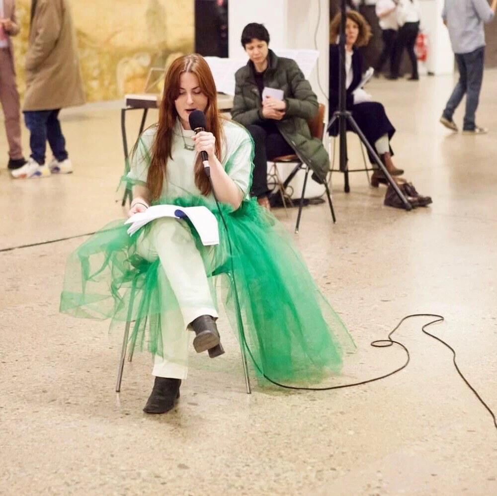 Charlotte Gash seated in a green tulle costume, reading into a microphone in a gallery.
