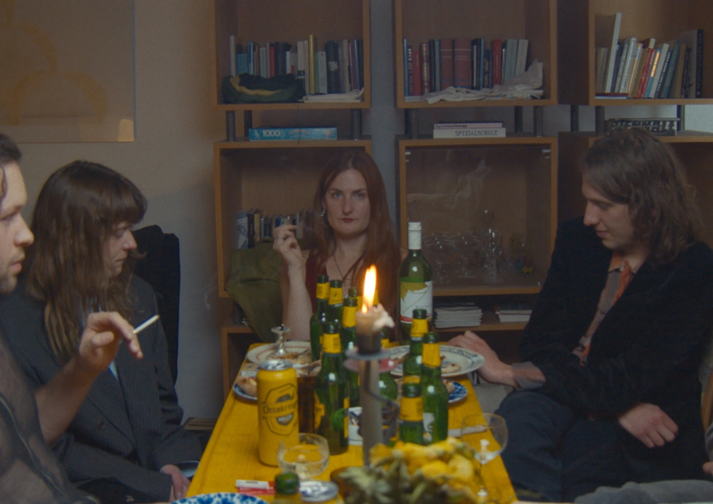Friends gathered around a yellow table with beer bottles and a lit candle in front of bookshelves.