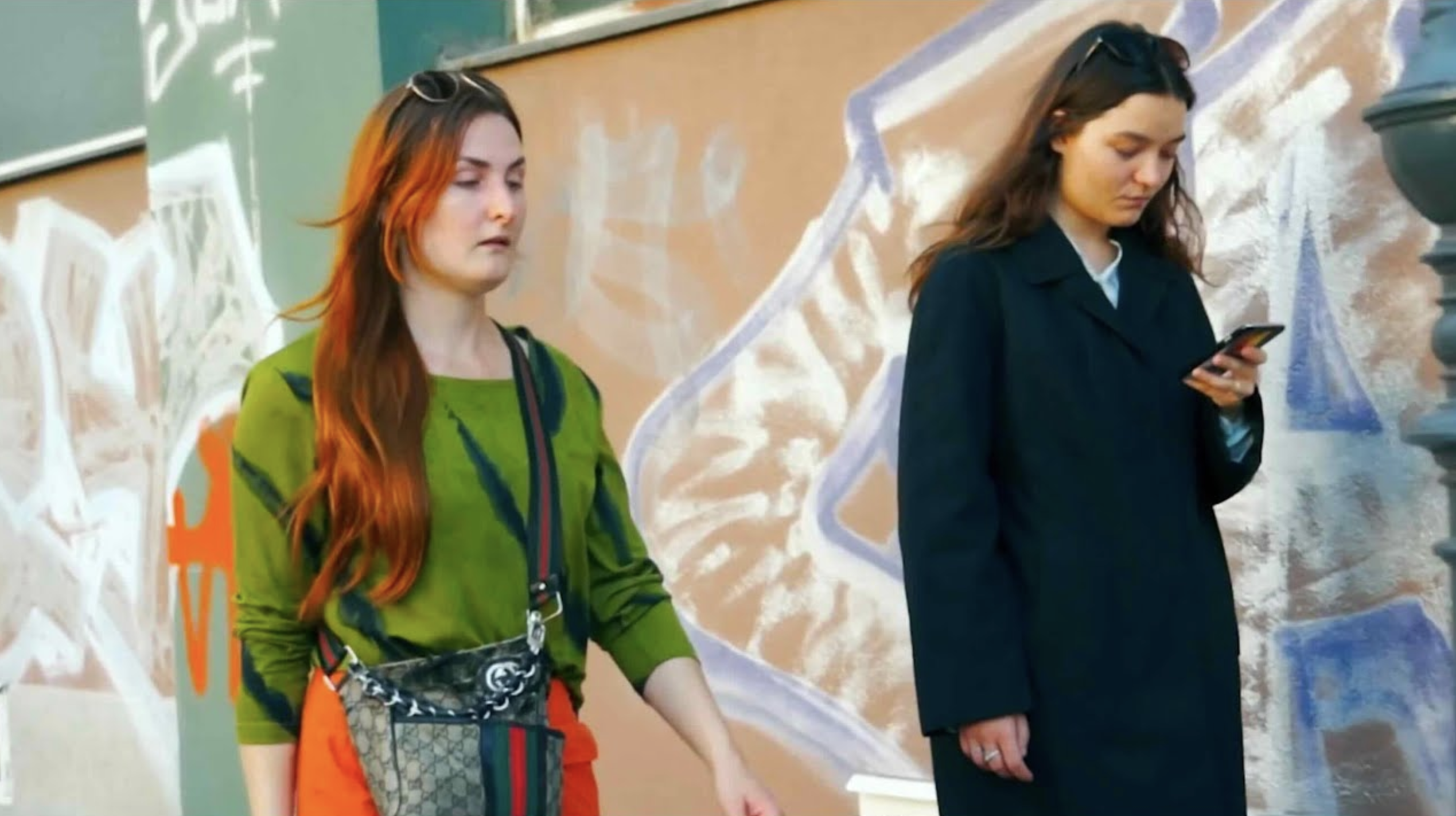 Two women standing in front of a painted wall, one in a green top and one in a dark coat holding a phone.