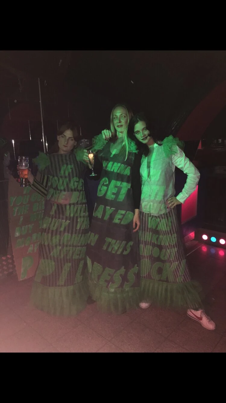 Three performers wearing dresses with projected green lettering in a club.