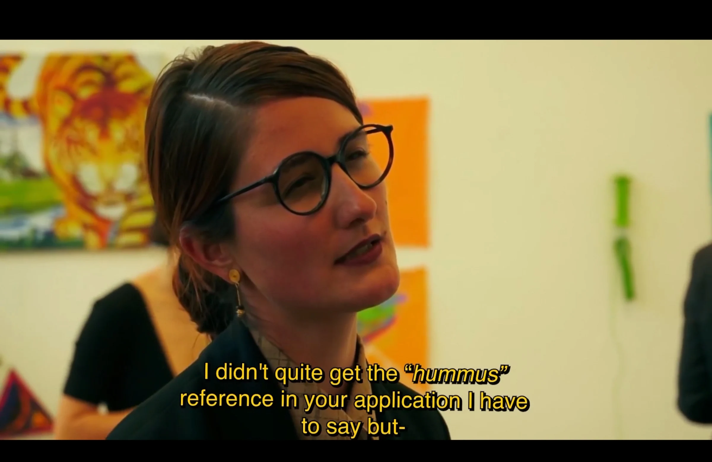 A character wearing glasses at an exhibition, discussing the hummus reference in an application.