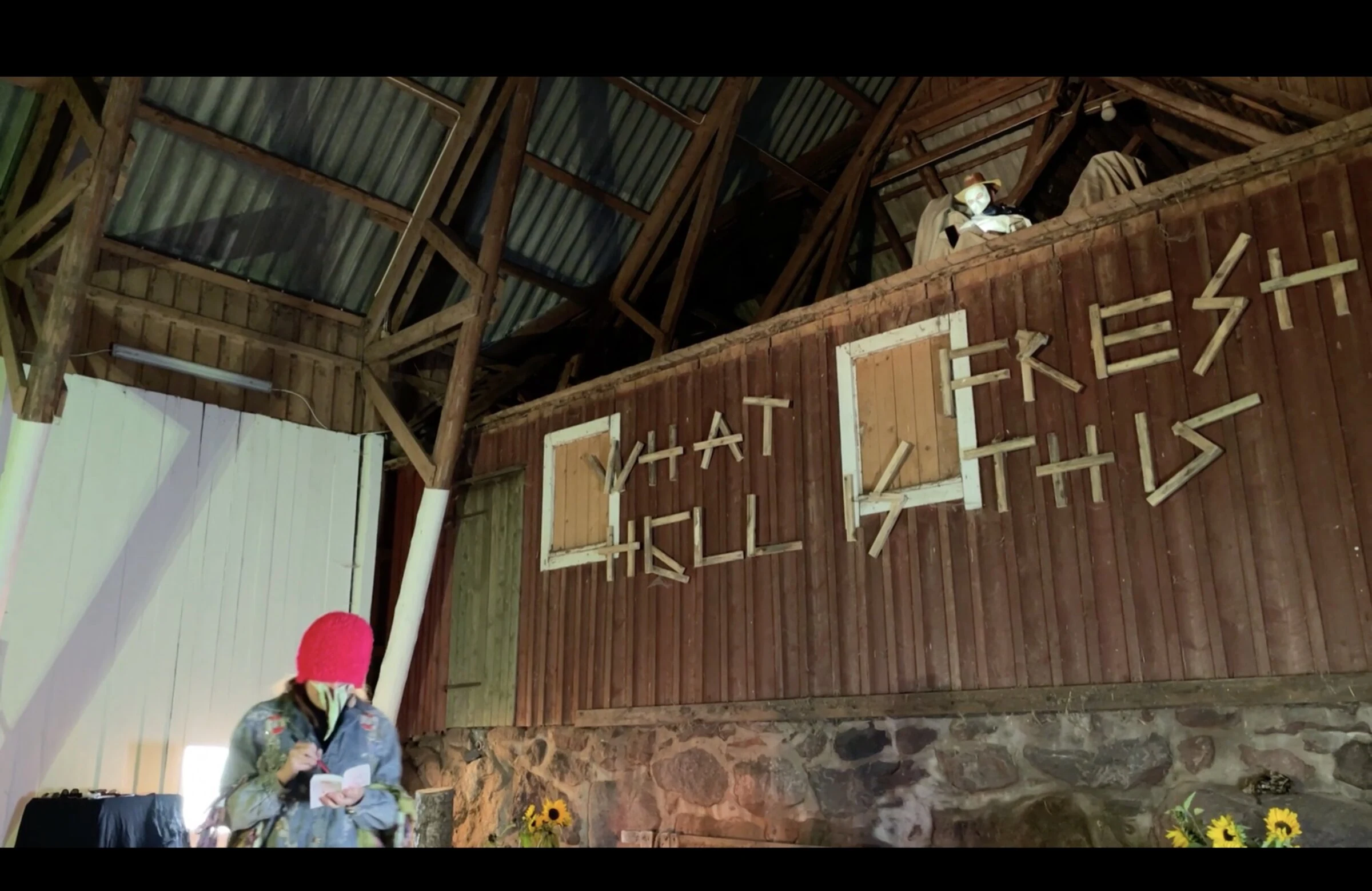 A performer in a red hat reading beneath another performer in the barn loft.
