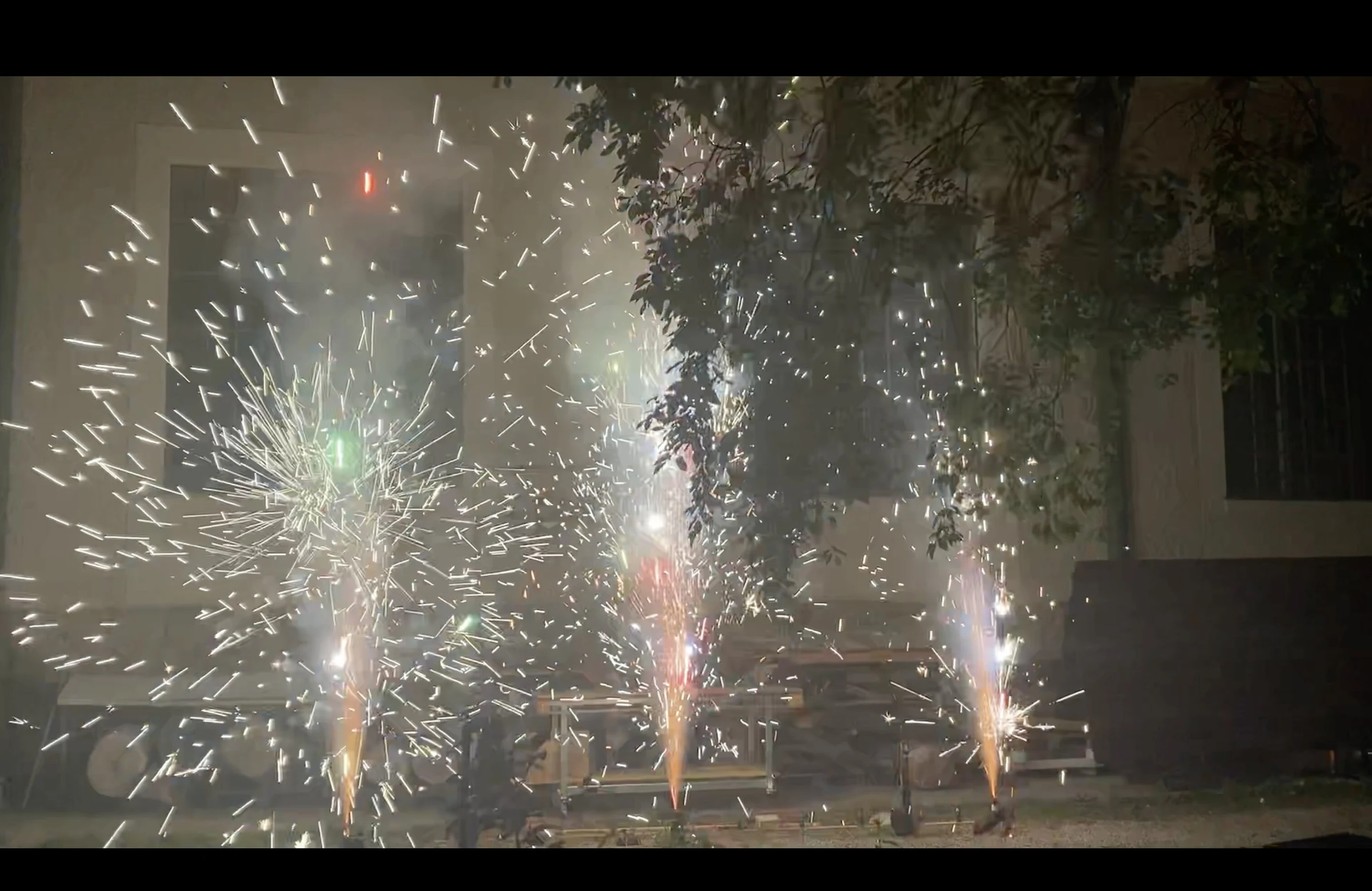 Three fountains of white sparks spraying across the courtyard beneath tree branches.