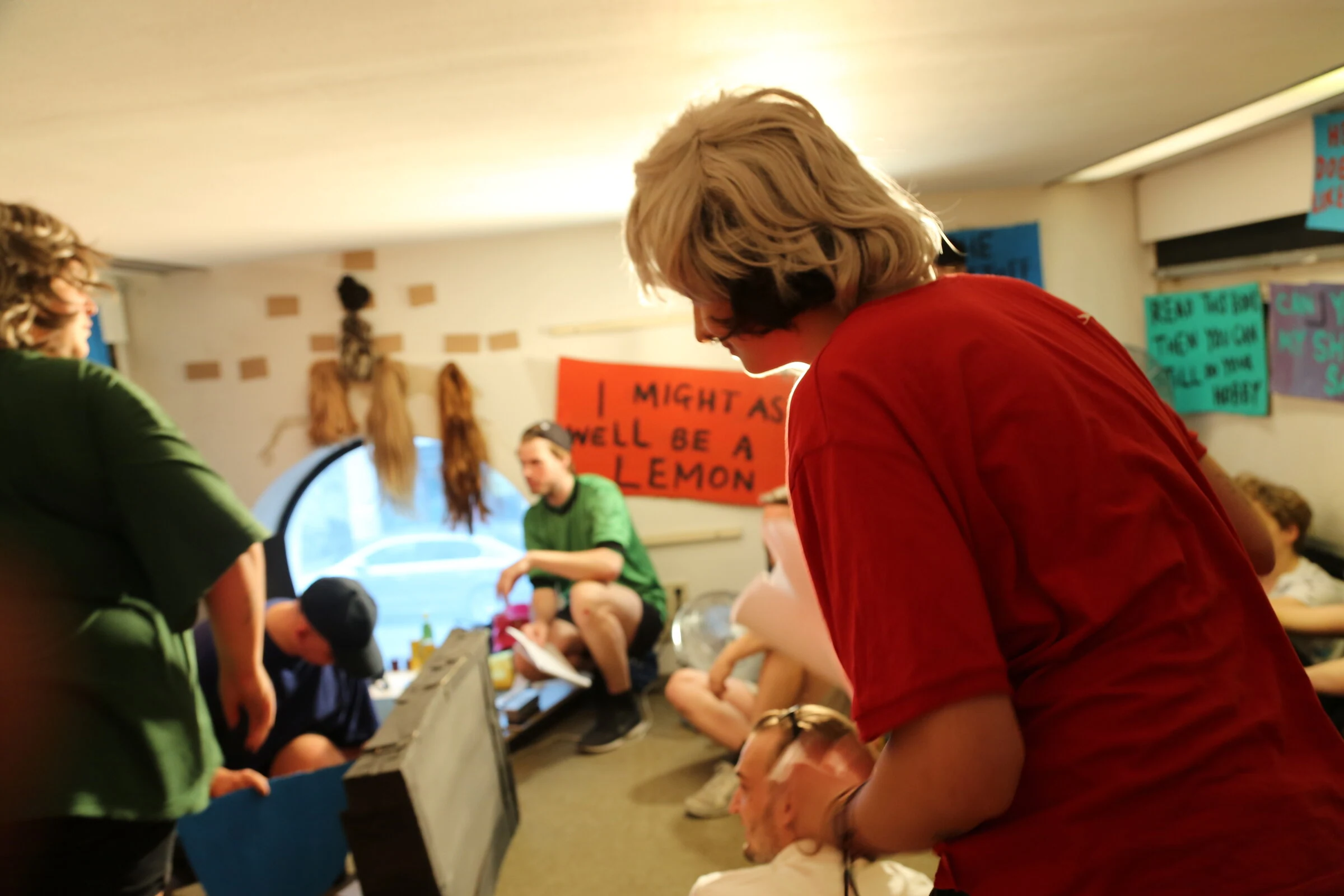 A performer in a blond wig and red shirt leaning forward among the performers and audience.