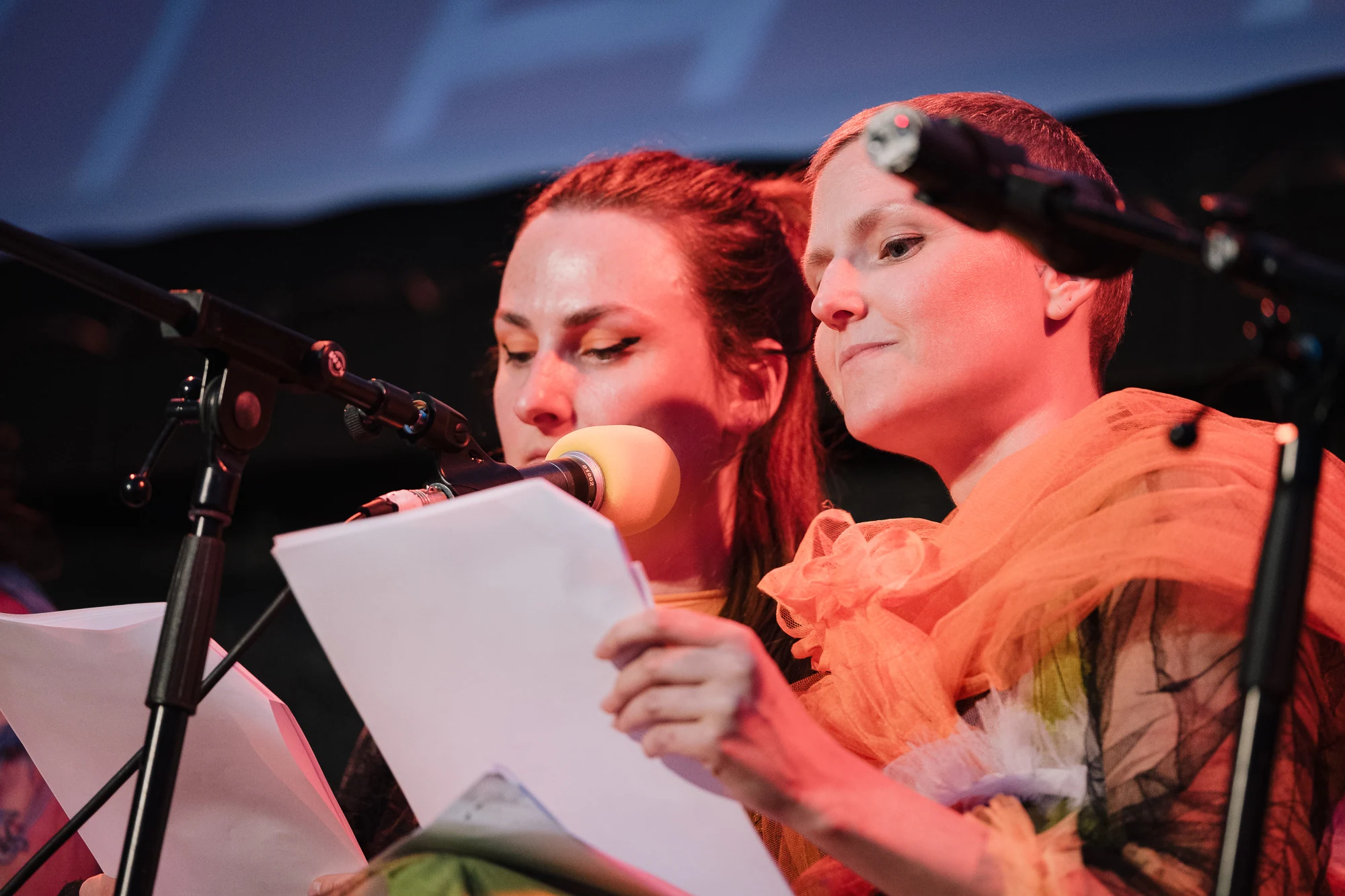 Two performers sharing a script beside microphones, one wearing an orange tulle collar.