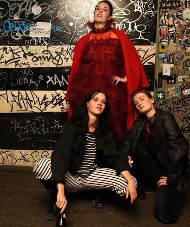 Three performers posing against a graffiti-covered wall, one in a burgundy tulle costume.