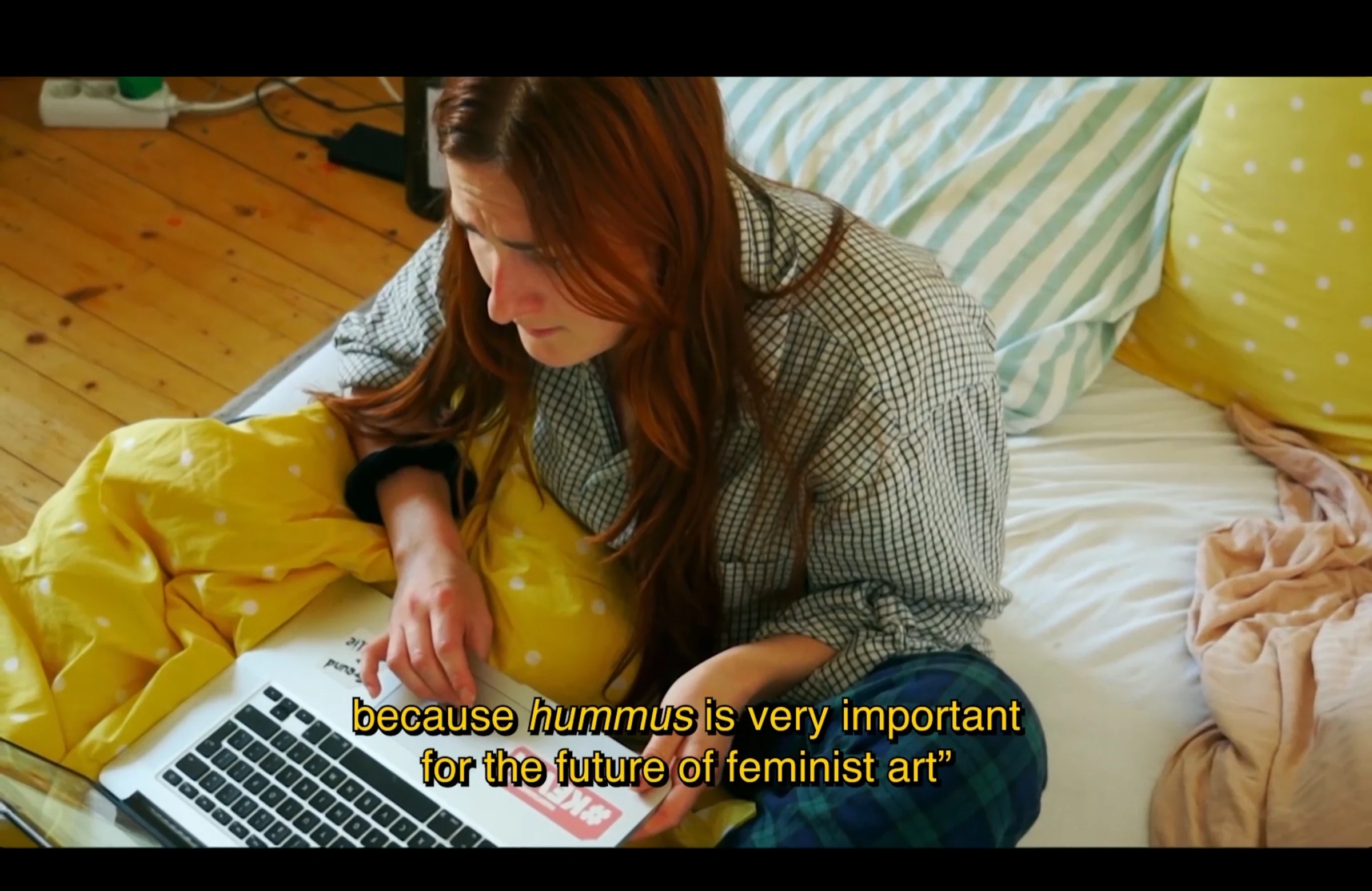 A character using a laptop on a bed with yellow bedding, discussing hummus and feminist art.